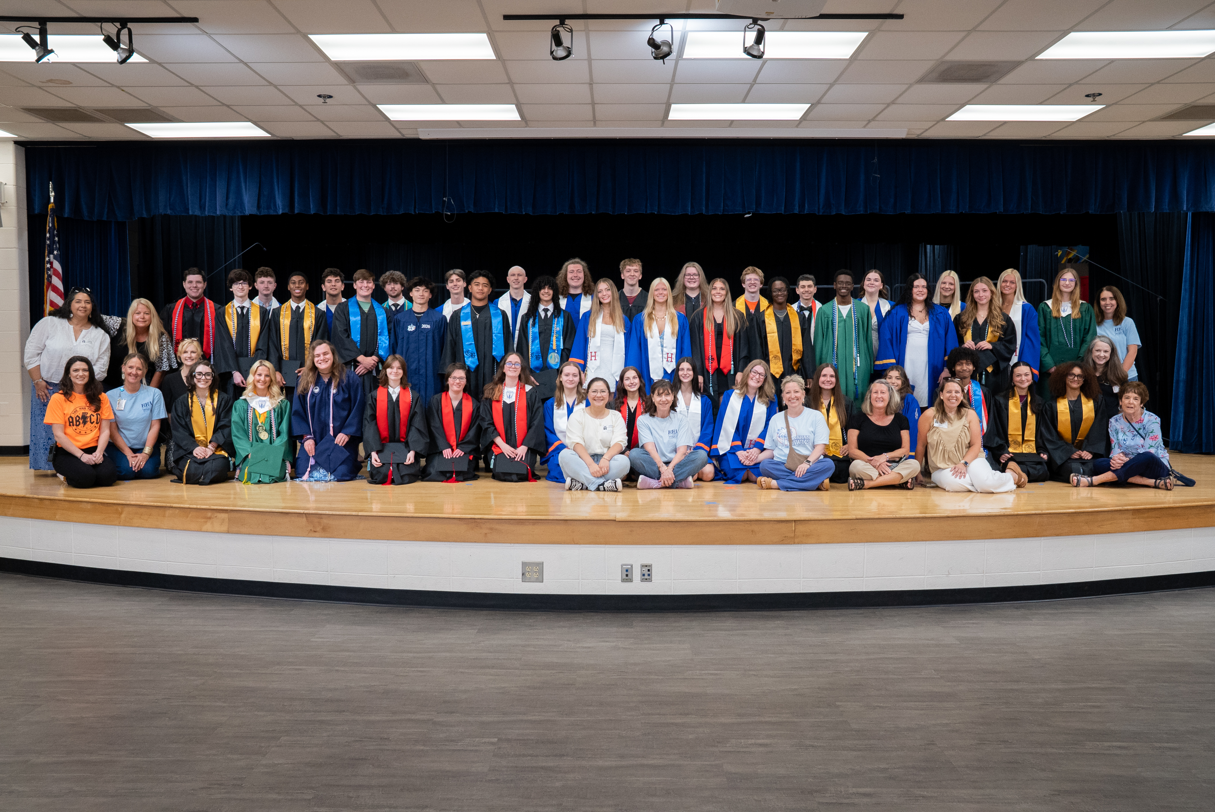 A group of people in gowns and caps pose for a picture on a stage with a blue curtain in the background.