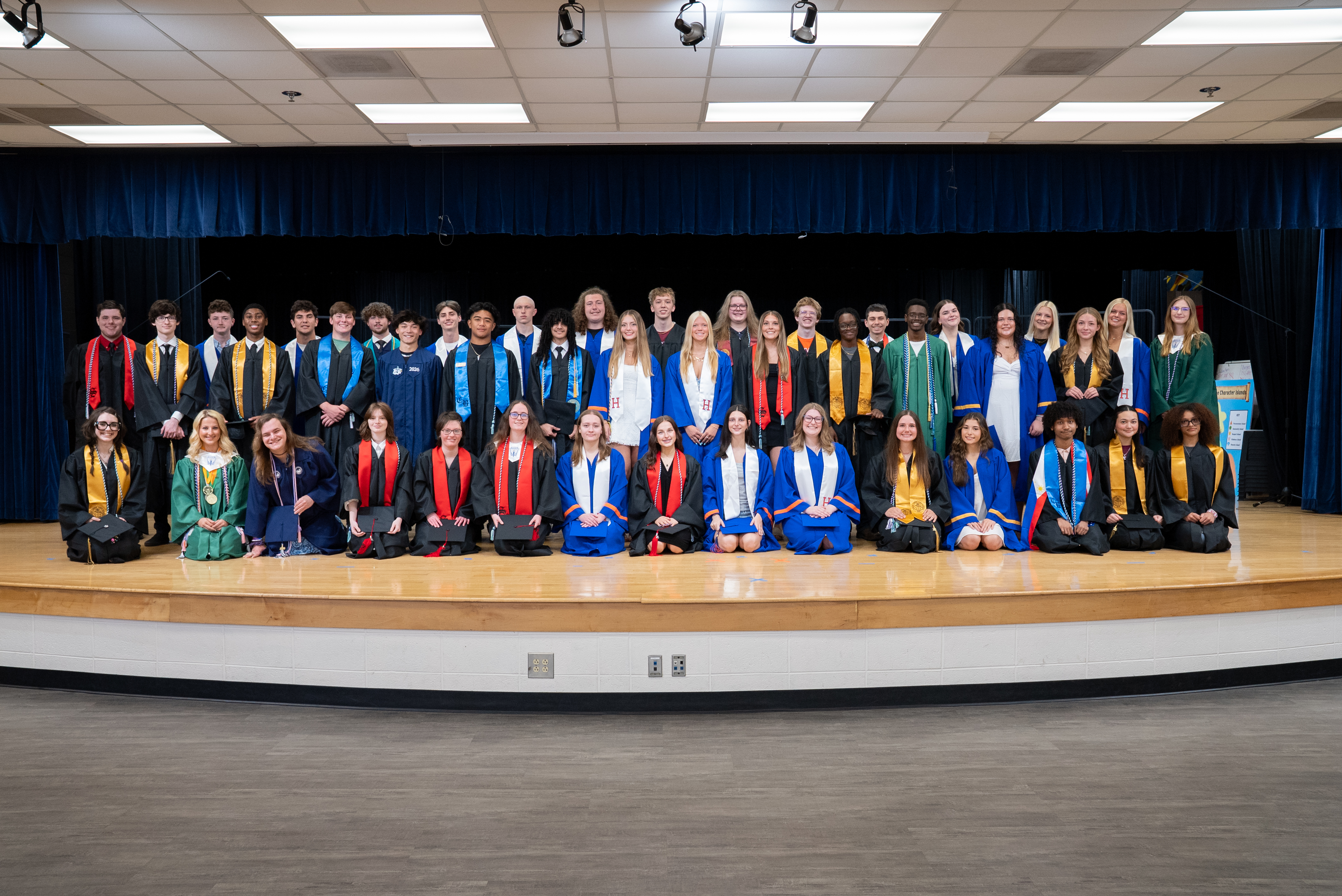 A group of graduates in various colors of gowns and stoles, standing on a stage.