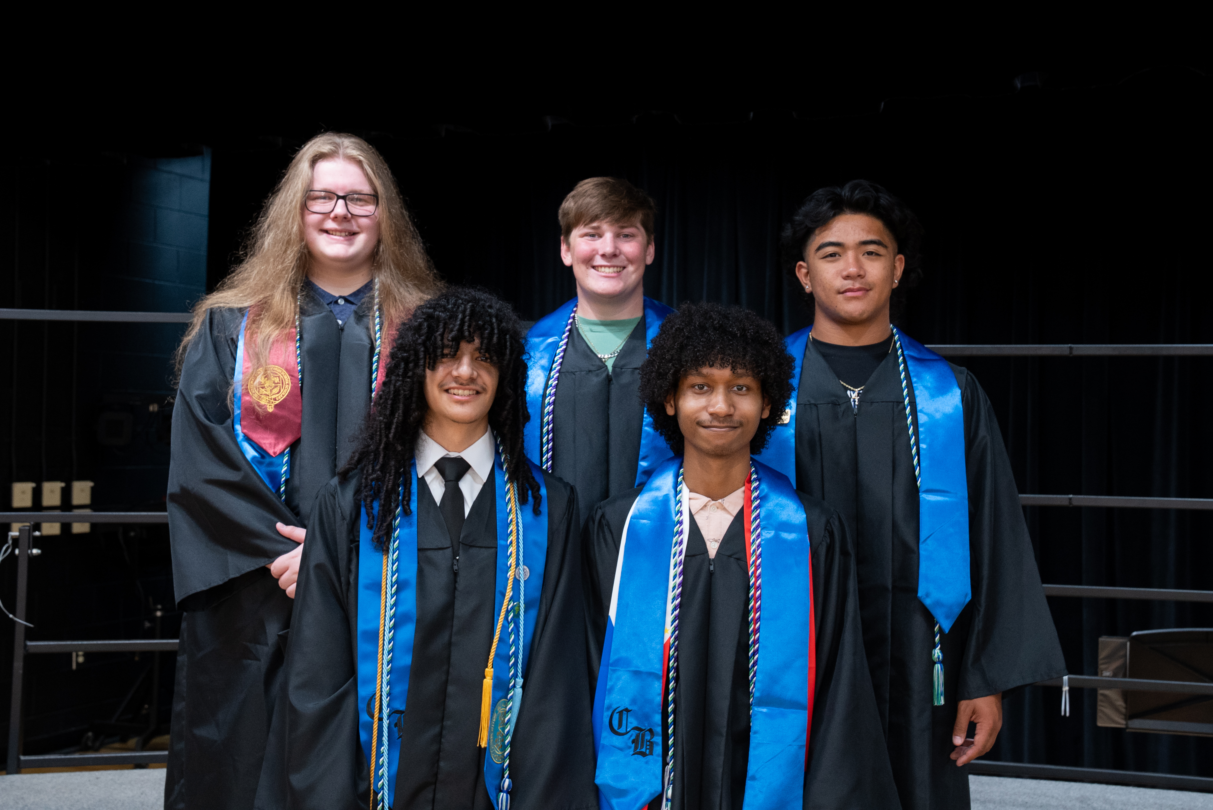 Five people in graduation robes and stoles pose for a photo in a theater setting.