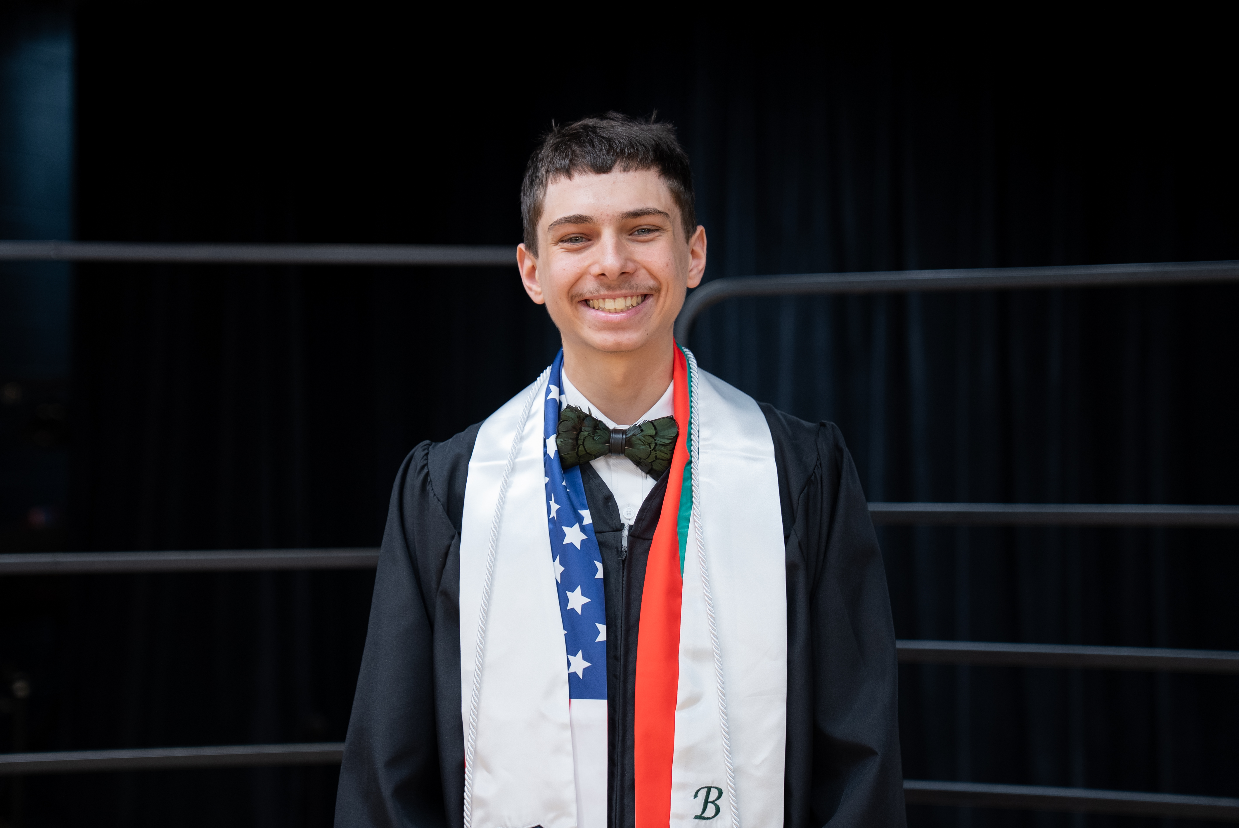 A young man in a black graduation gown, a bow tie, and a white stole with the American flag design.