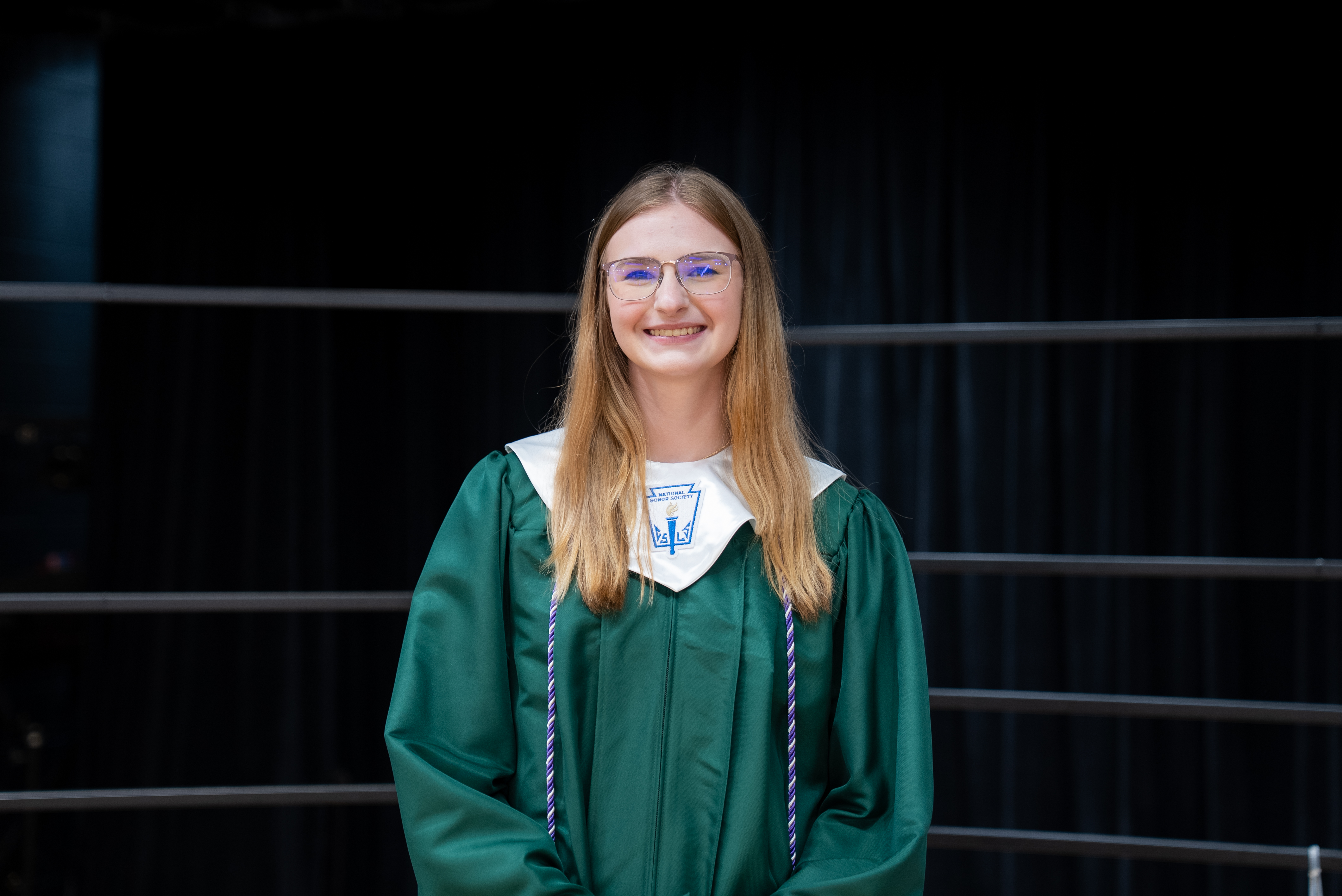 A person in a green graduation gown with white trim stands in front of a black curtain.