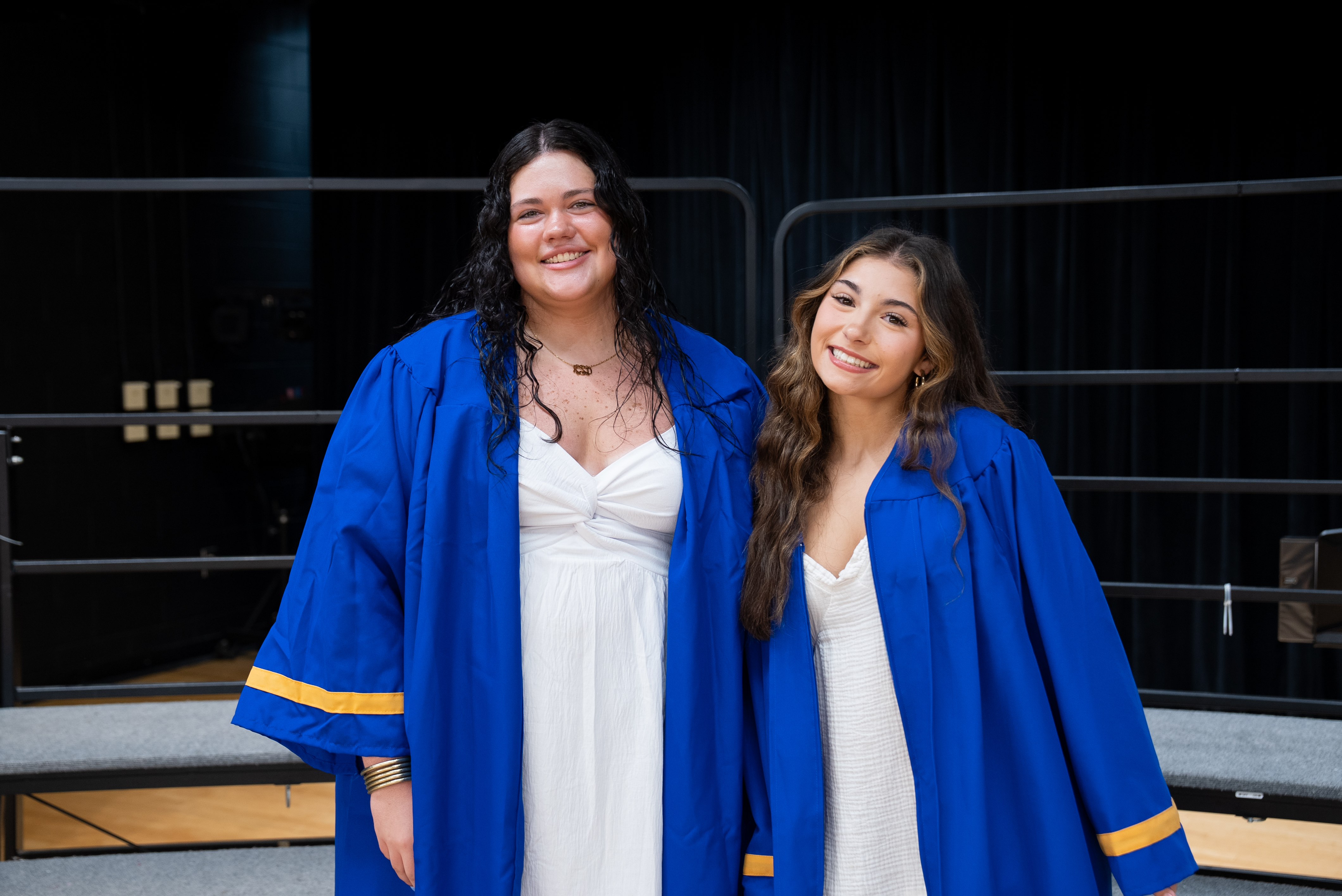 Two women in blue graduation robes stand next to each other. One wears a white dress, and the other has a necklace. Behind them is a black curtain and metal railings.