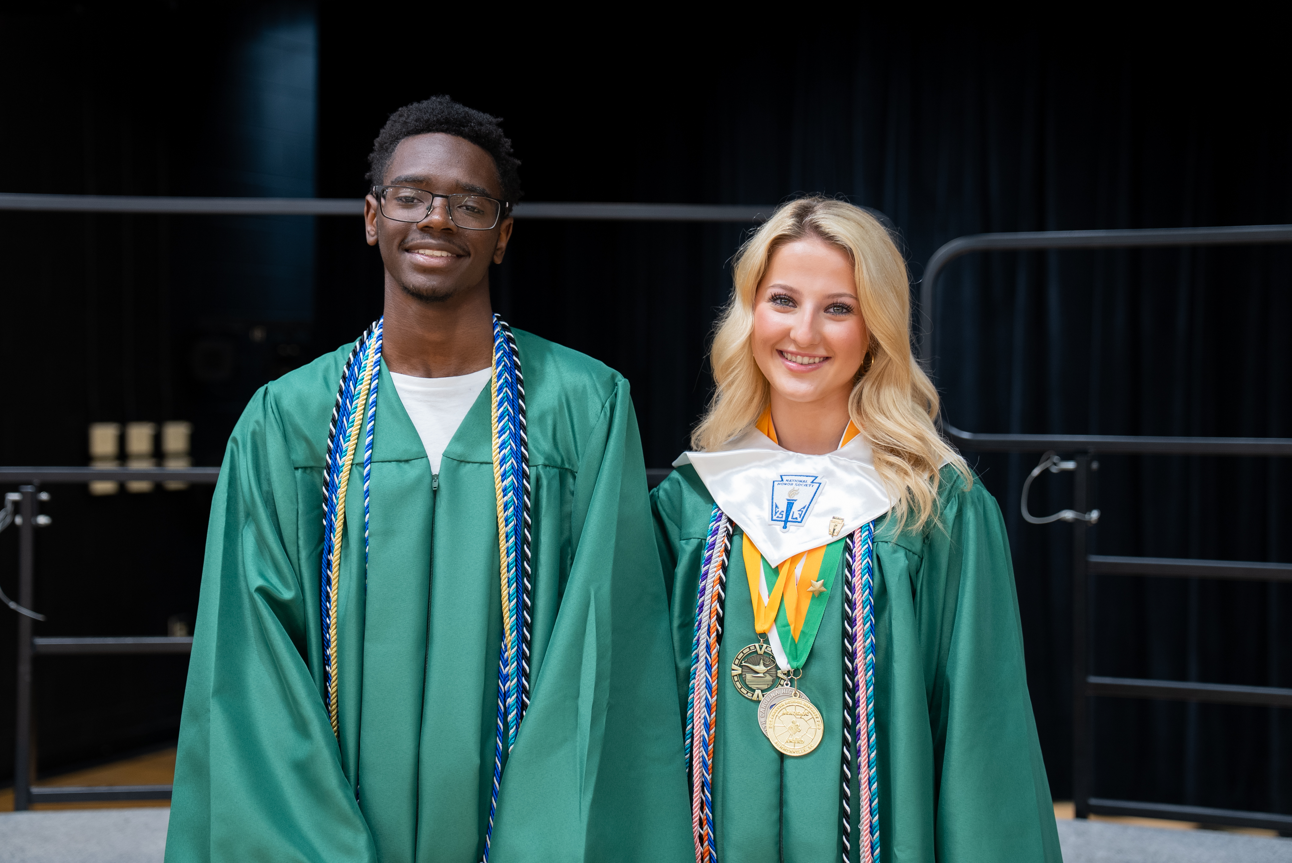Two individuals in green graduation robes, one with a medal, pose for a photo in a theater.