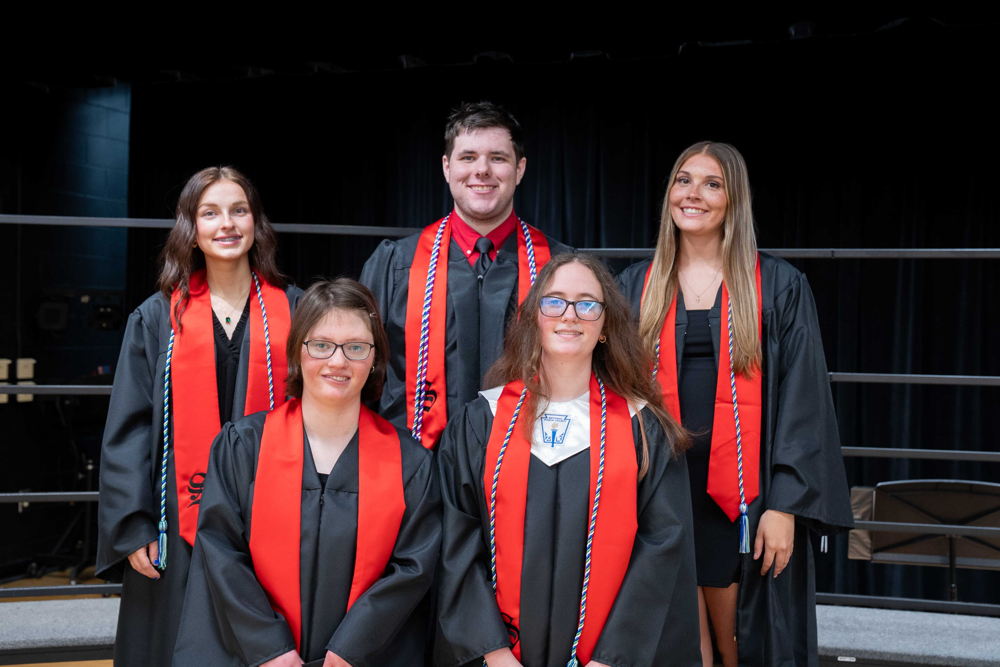 Five individuals, possibly graduates, dressed in black gowns with red sashes, stand together in a row on a stage.