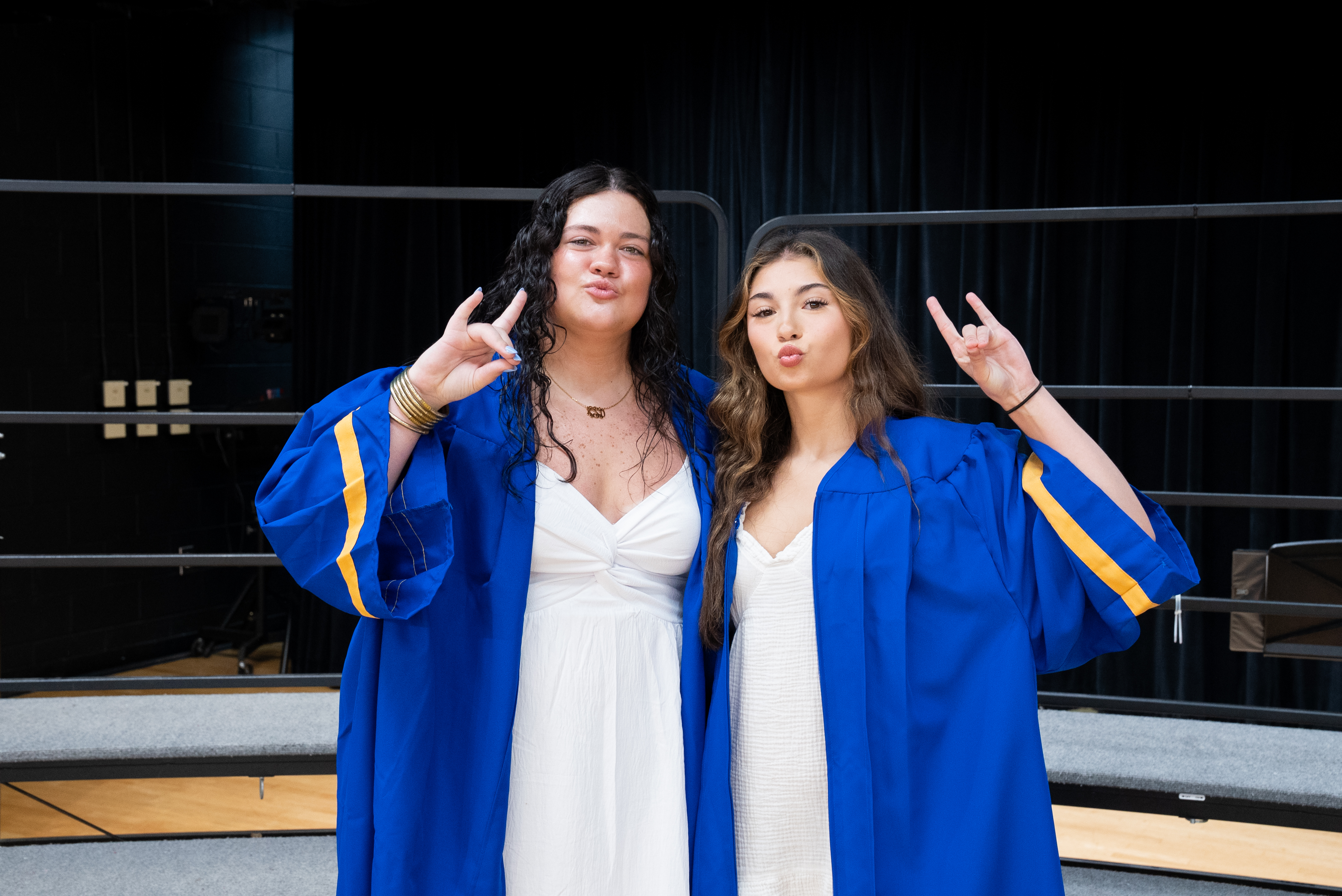 Two women in blue graduation gowns make peace signs in a school auditorium with a black backdrop.