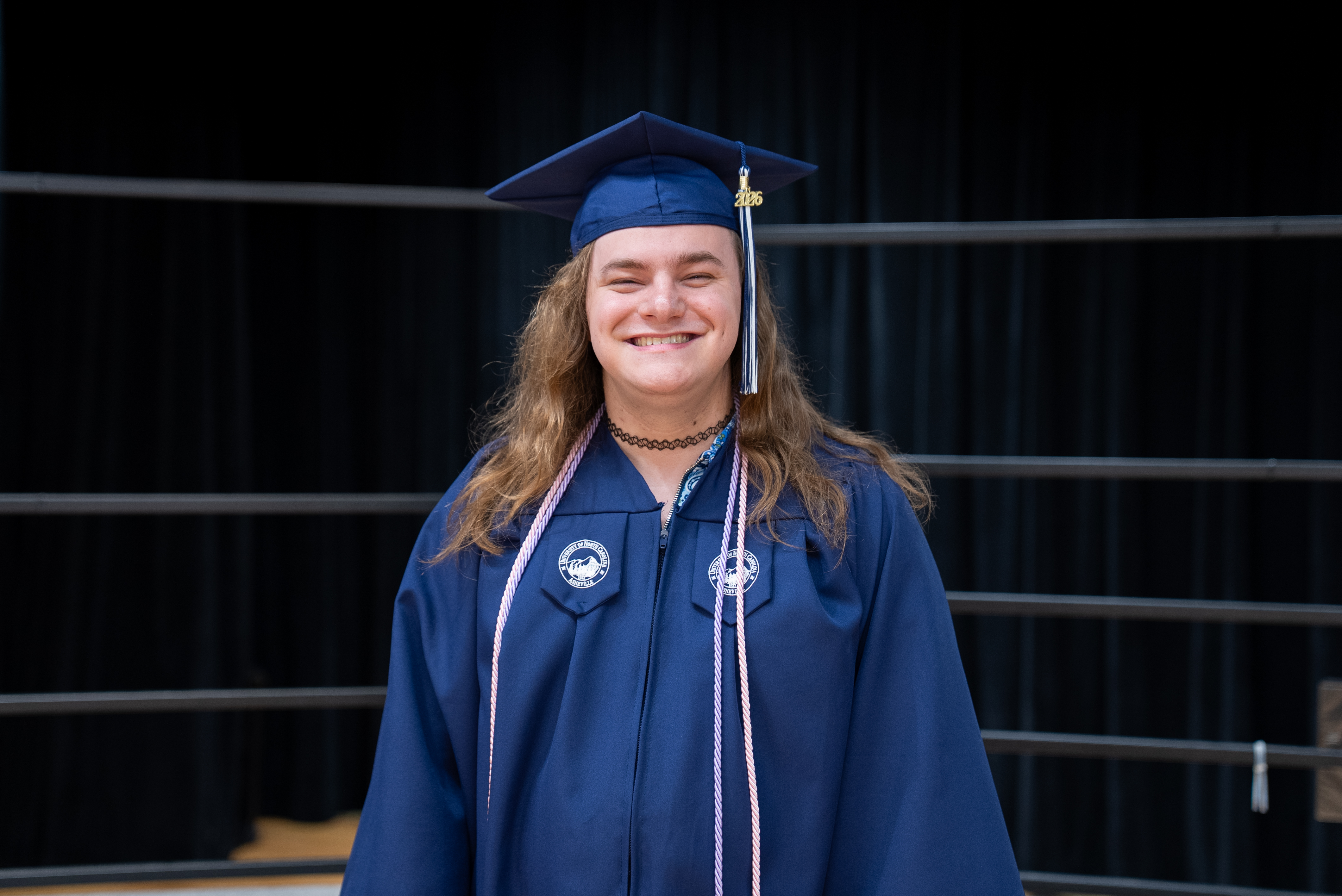 A person in a blue graduation gown and cap stands smiling in front of a black curtain.