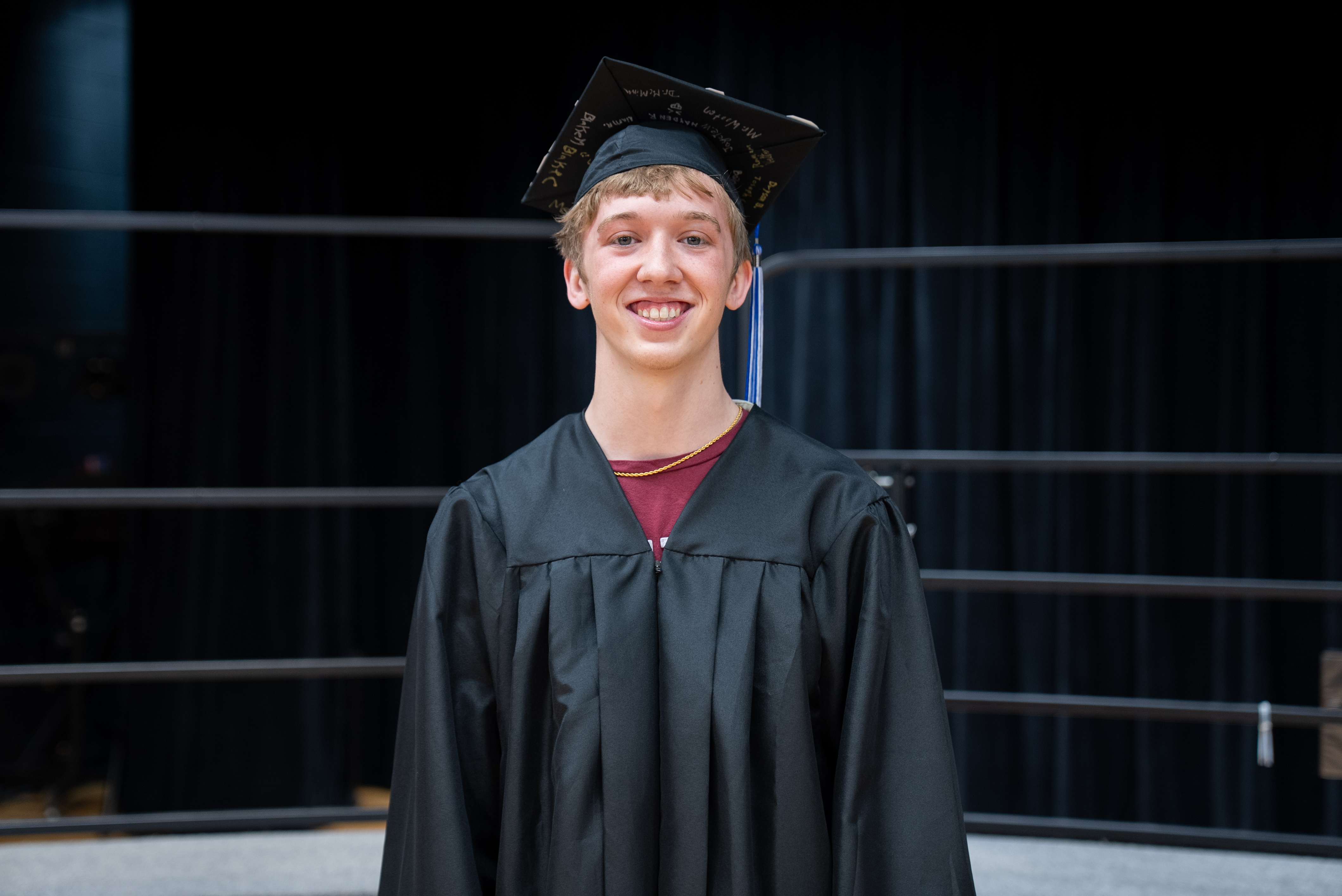 A man in a black gown and mortarboard stands, smiling, with a black curtain background.
