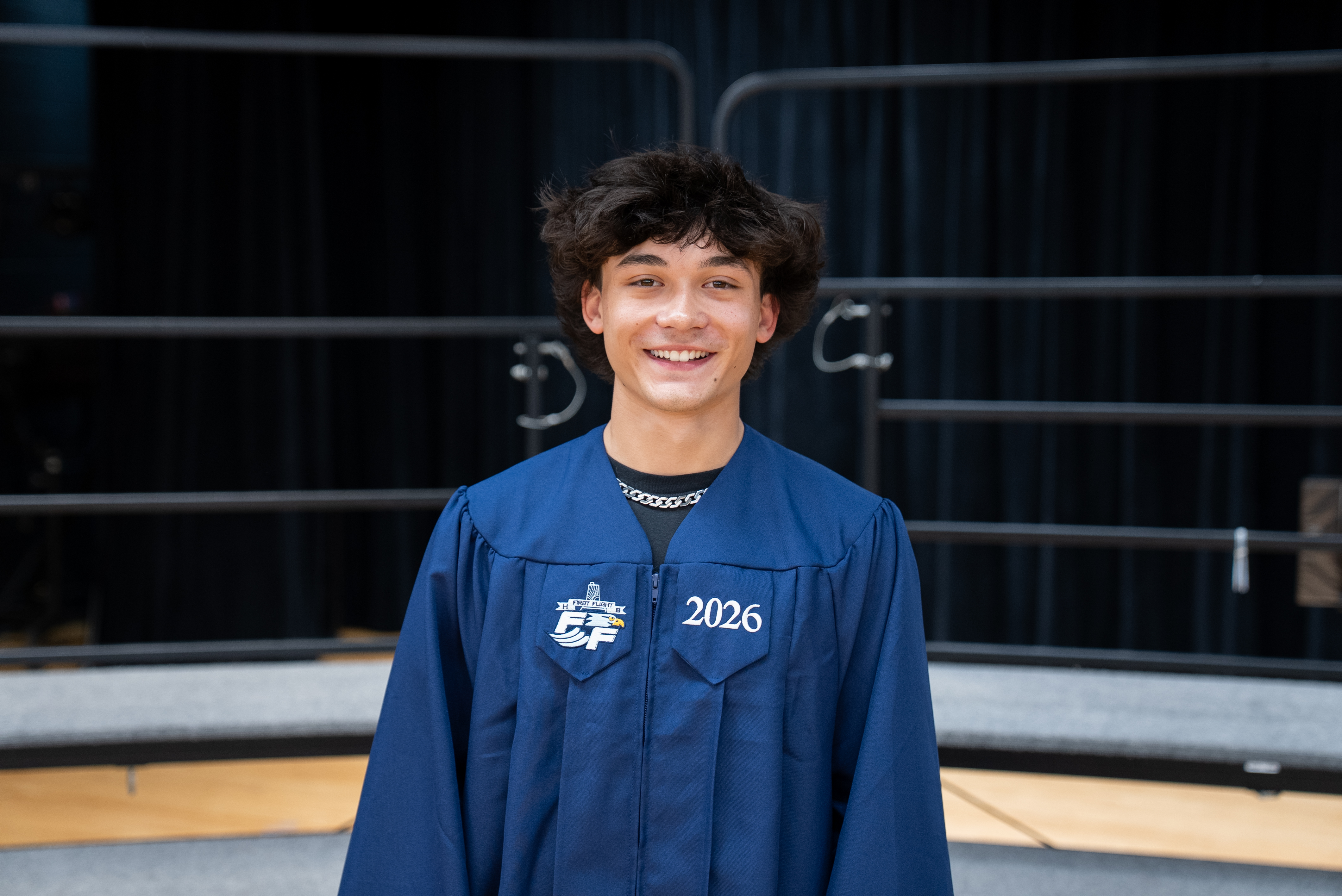 A young man in a blue graduation gown smiles and poses for a photo on a stage.