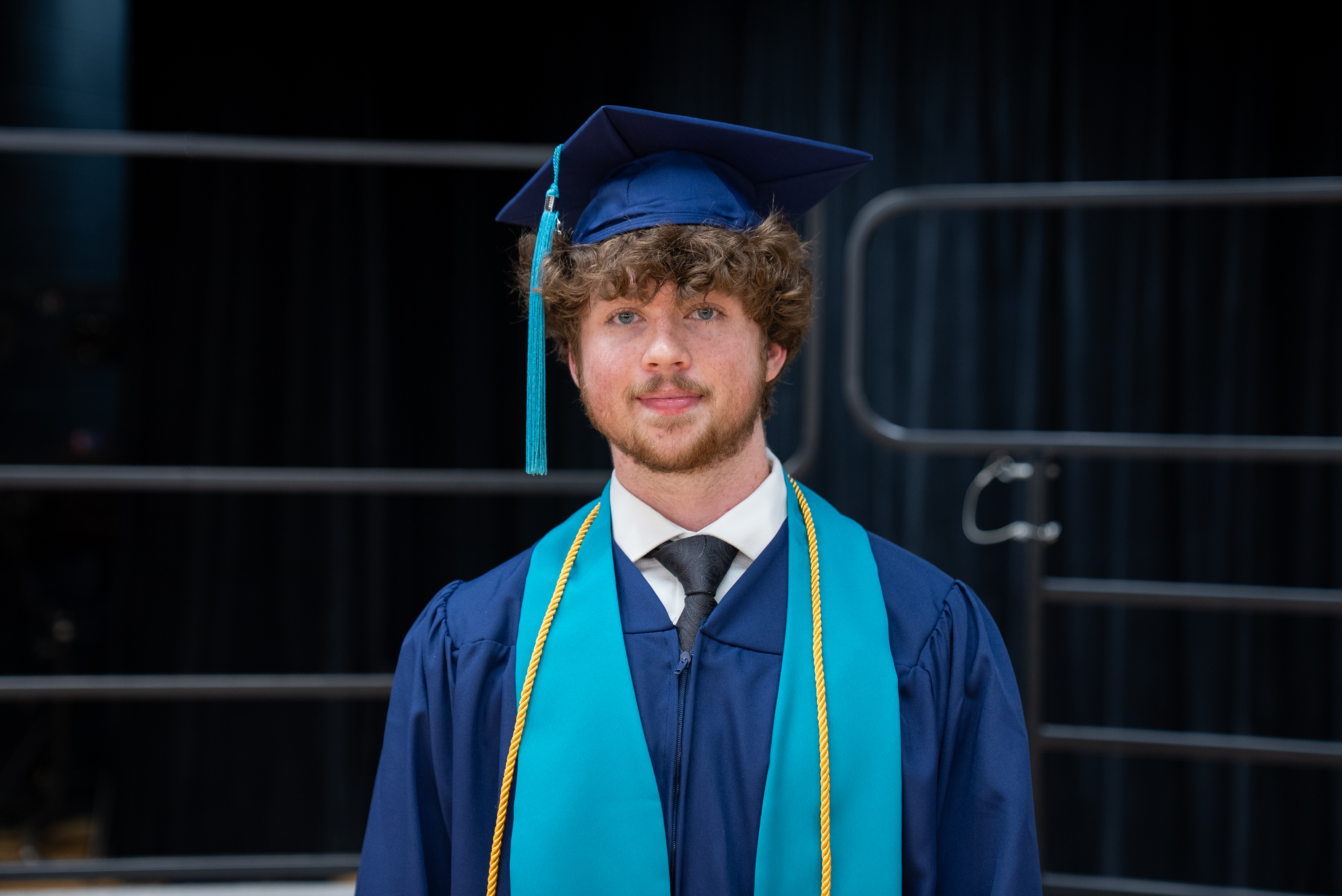 Man in cap and gown, with blue stole, stands in front of metal railings.