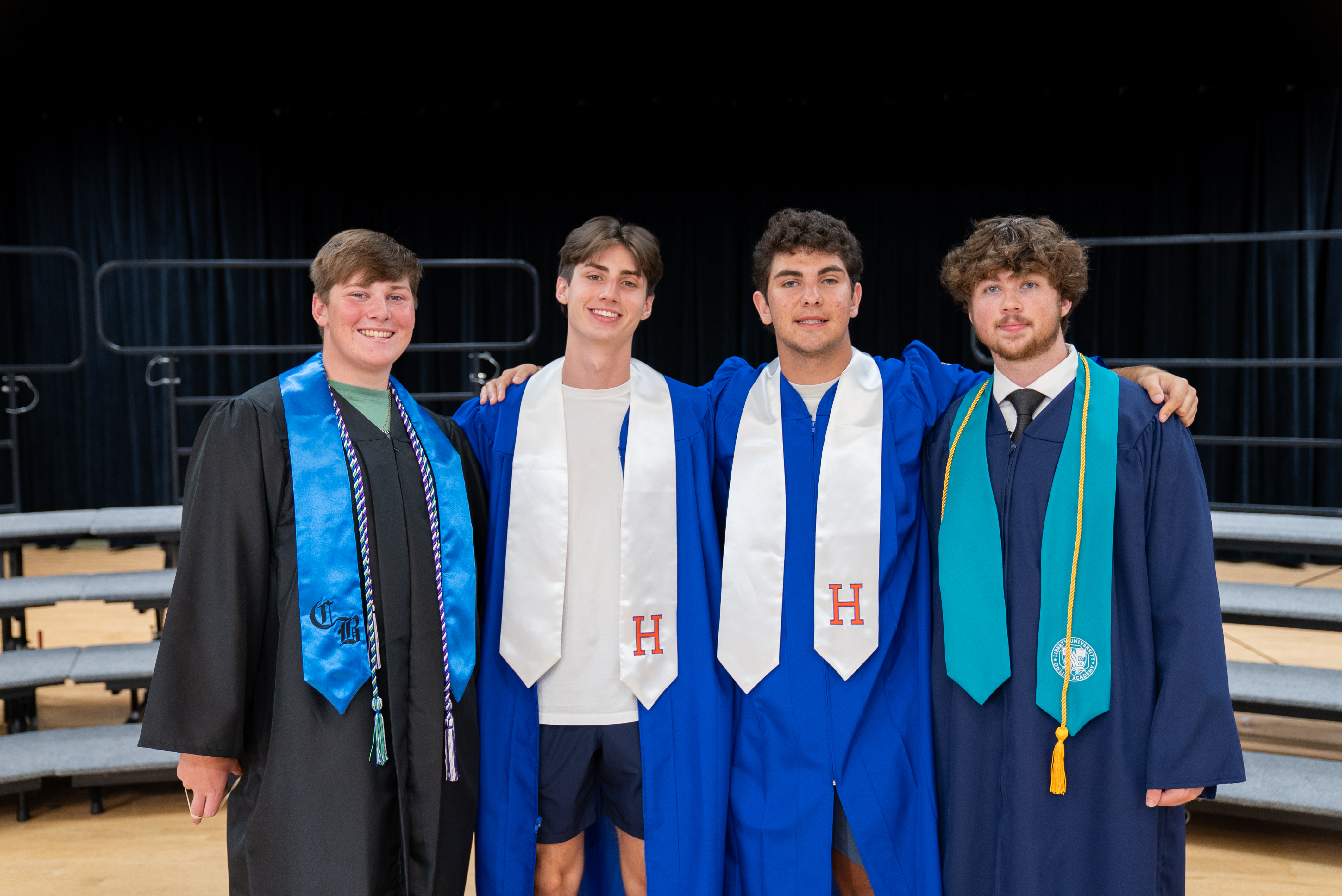 Four men in academic gowns stand together, three with arms around each other, on a stage with bleachers.