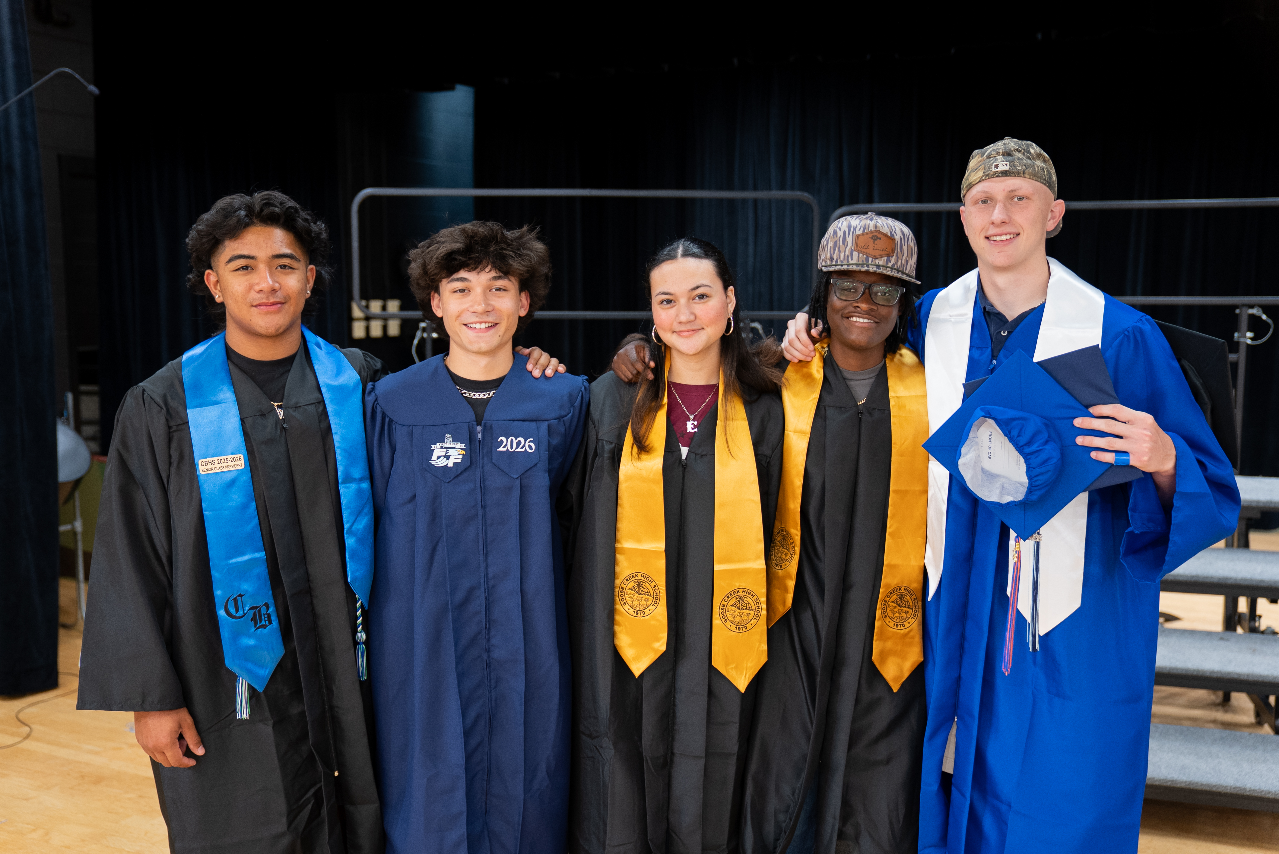 Five people in academic robes and caps pose for a photo on a stage with stairs.