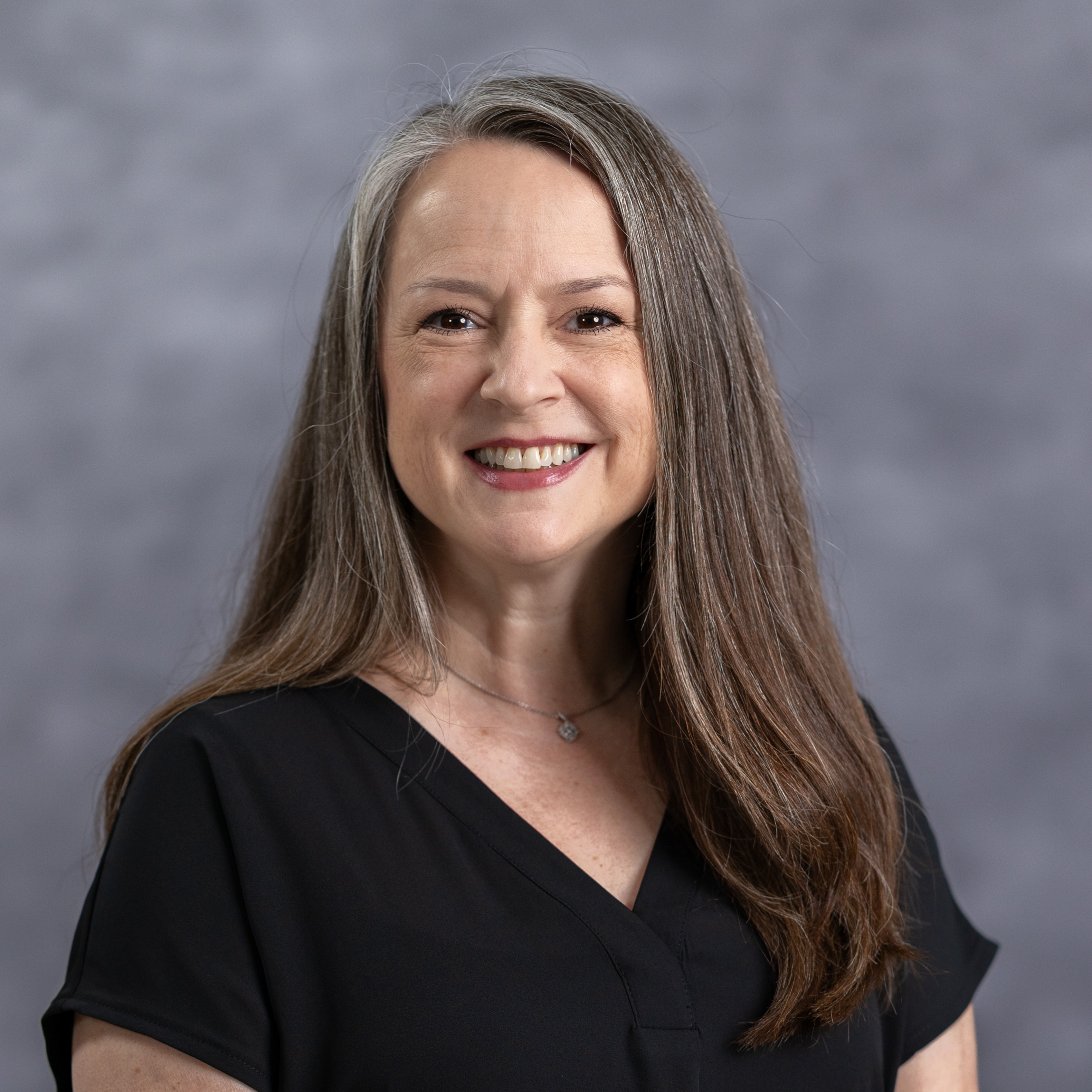 An adult woman with long brown hair, wearing a black shirt, smiles and poses for a photo.