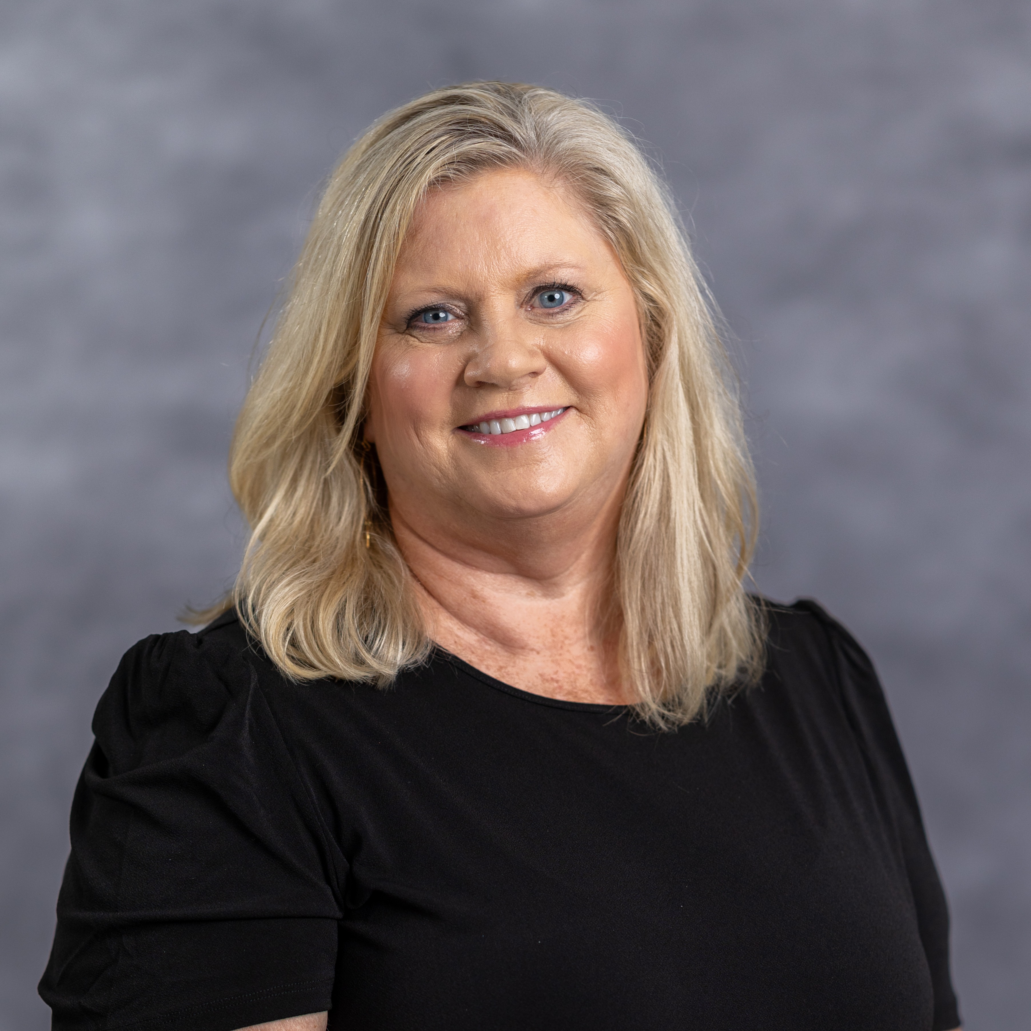 A woman with blonde hair smiles, wearing a black shirt. Her hands rest on her hips. Background is solid gray.