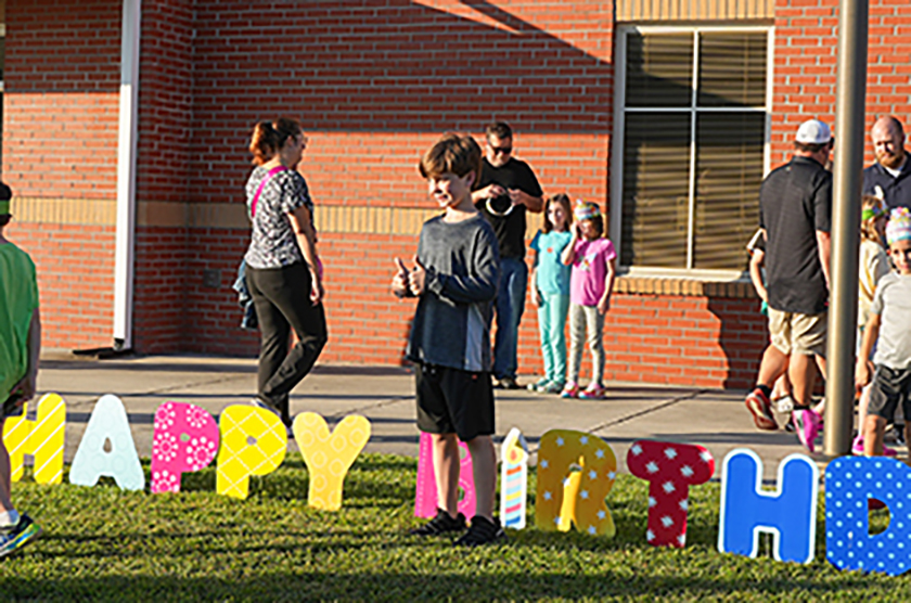 People in front of a brick building. Letters spell "Happy Birthday" on grass. Candles on top of "Happy Birthday" letters.