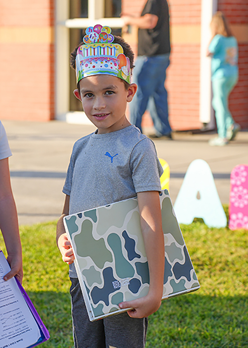 A young boy with a birthday crown holds a camouflage backpack in a grassy area. A building is behind him.