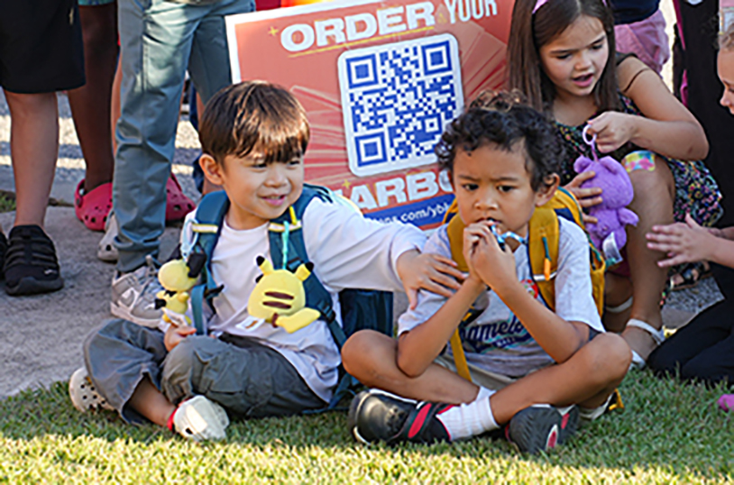 Two young children sit on grass, one holds a toy, the other a snack. They wear backpacks. Behind them, a group of children and adults stand.