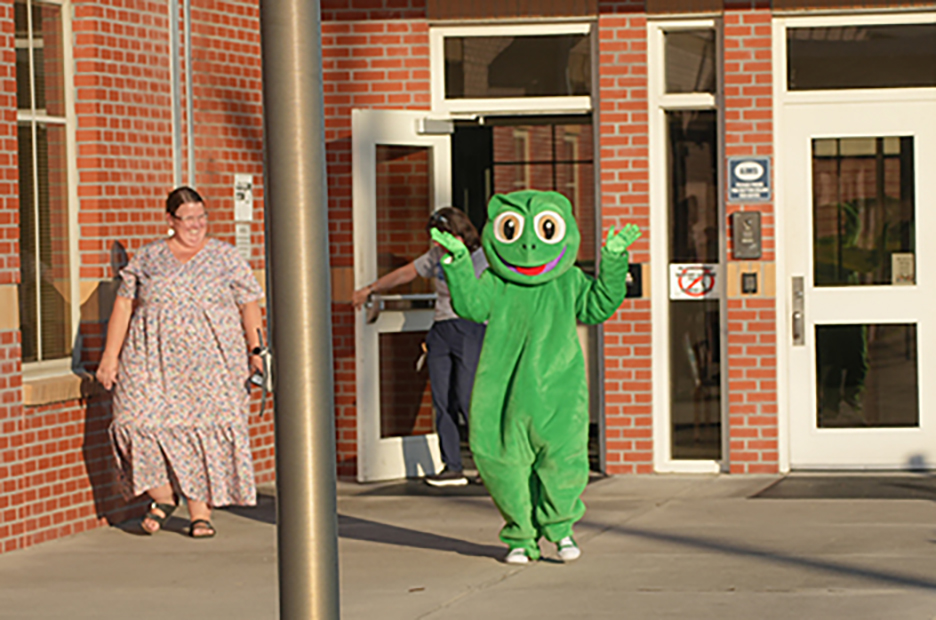 A person in a frog costume walks toward a building. A woman wearing a dress walks away.