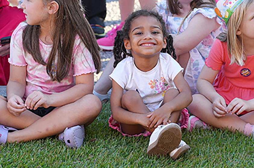 Children sit on grass, smiling. Two wear white tops, one in pink, another in orange. Background shows other children.