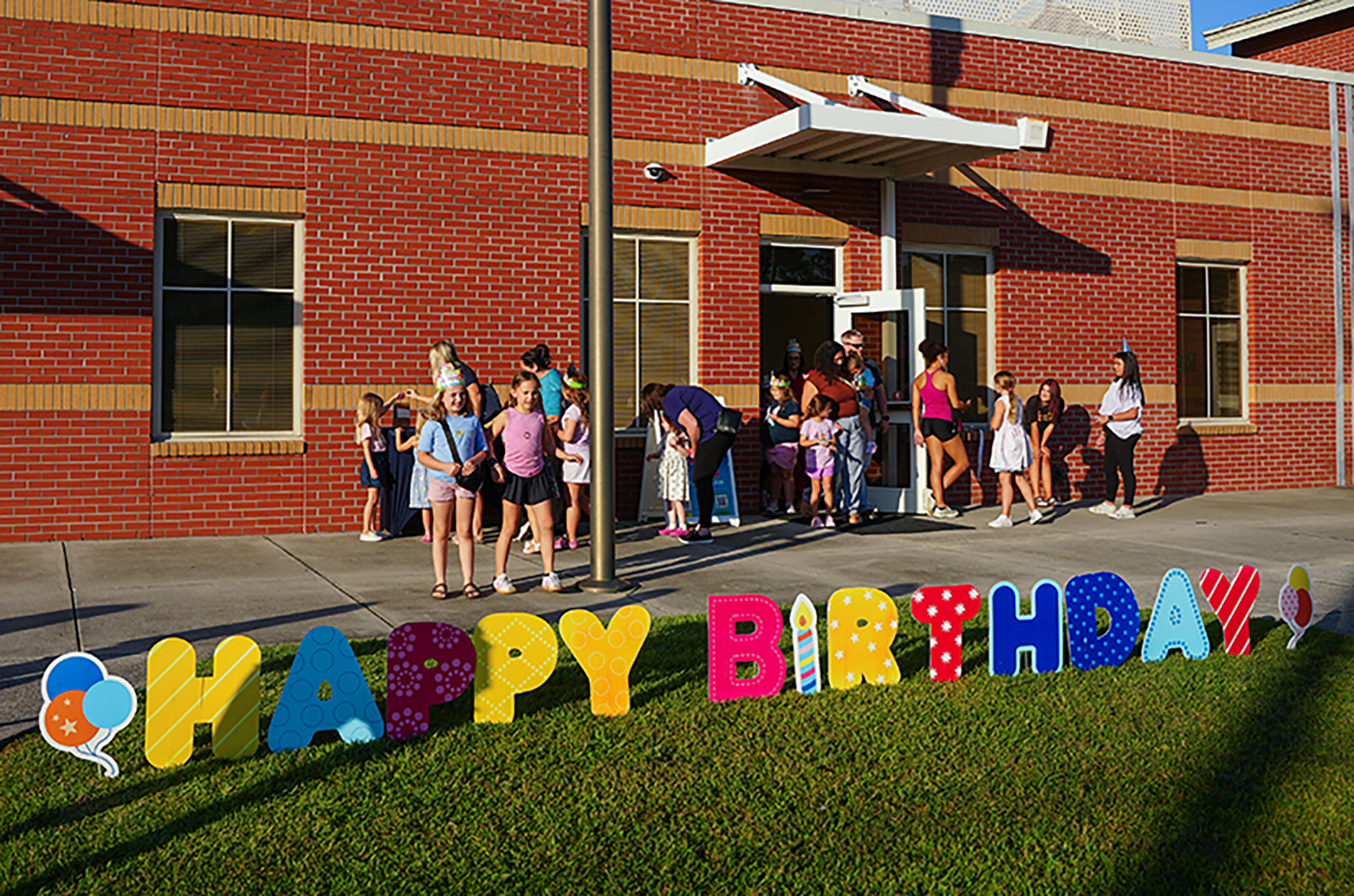 People gather in front of a red building with large "Happy Birthday" letters on the grass.