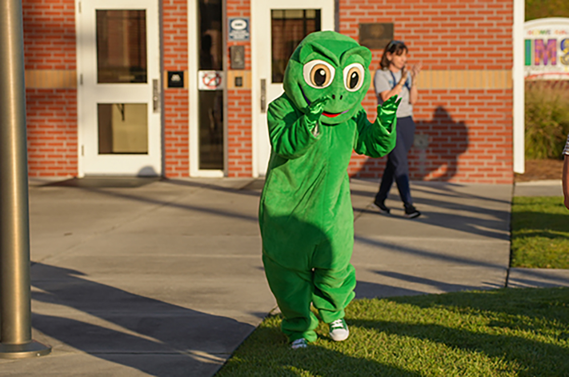 Person in green mascot costume waving near a building. A person in a gray shirt walks behind.