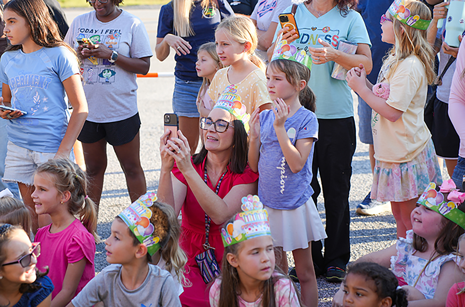 A group of children in party hats with a woman taking a photo with her phone.