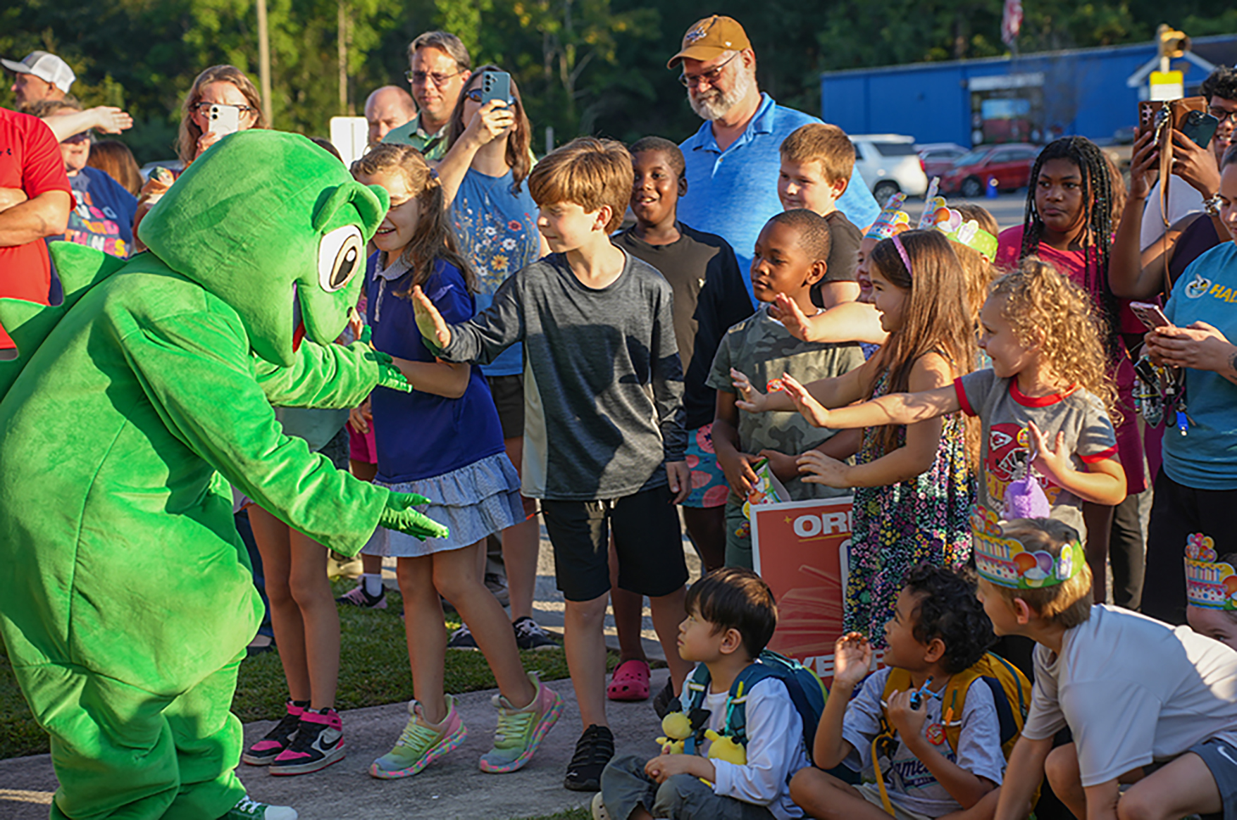 A mascot in a green suit interacts with children in an outdoor setting; several adults stand and crouch behind them.