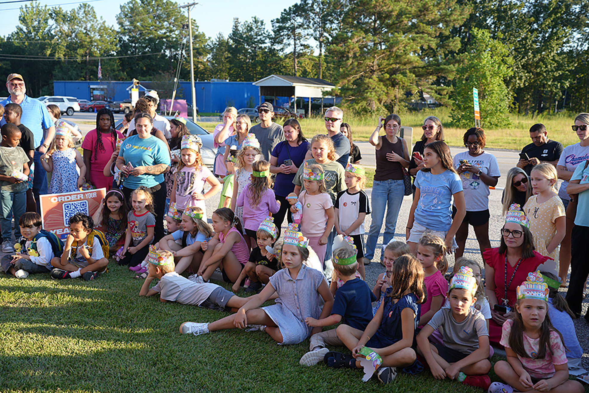 A group of people, including children, standing and sitting on grass. Many wear party hats. Trees and buildings in the background.