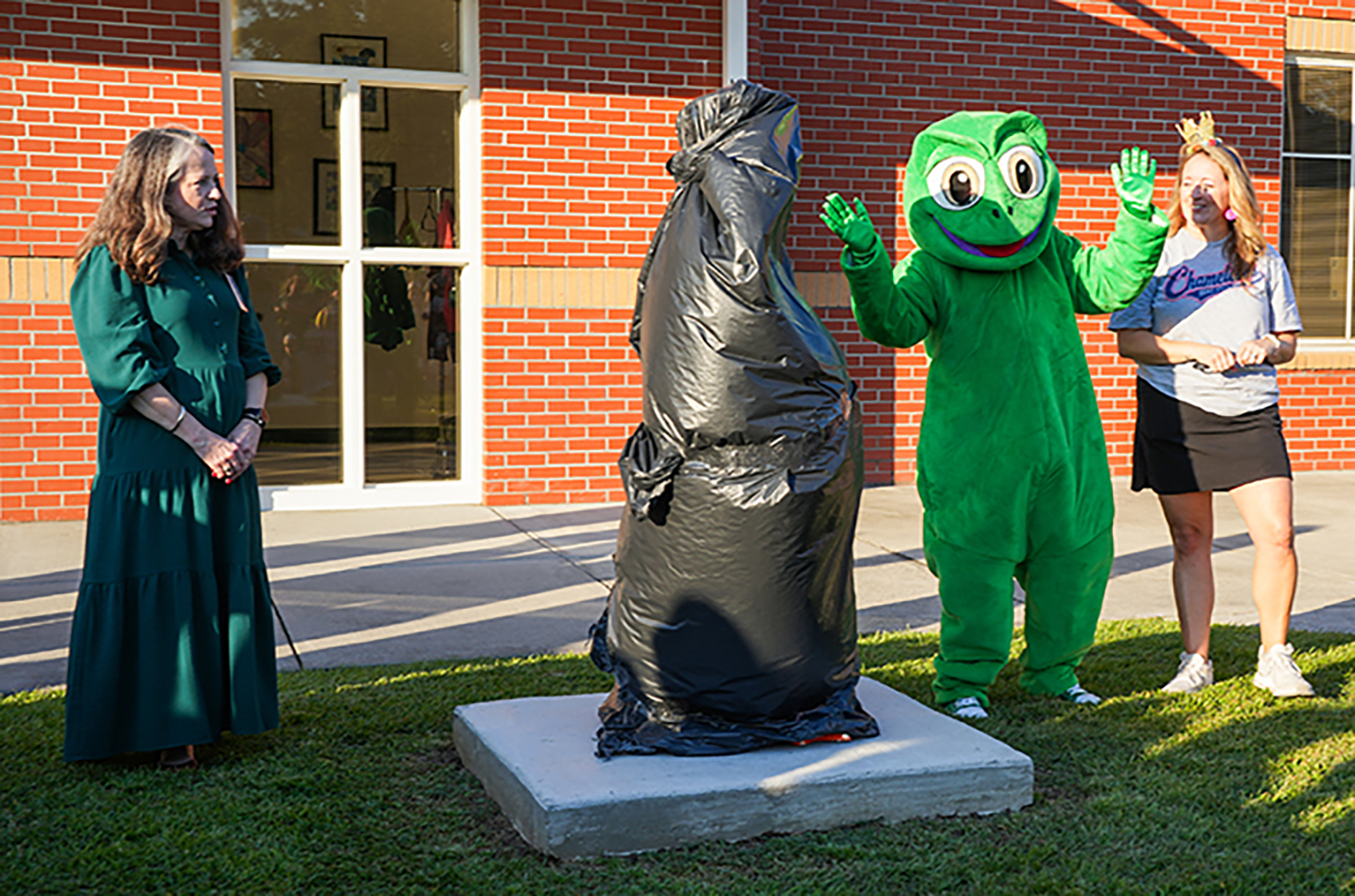 Three individuals stand near a sculpture. One wears a green mascot costume, another a black dress, and the third a white shirt and black skirt.