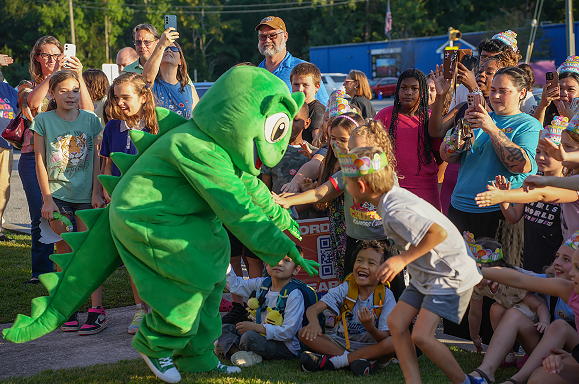 A green mascot entertains children in a park. Children smile and clap; one boy shakes the mascot's hand.