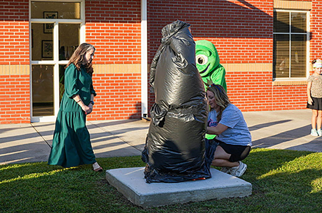 Two women prepare to unveil a wrapped sculpture in front of a brick building with large windows.