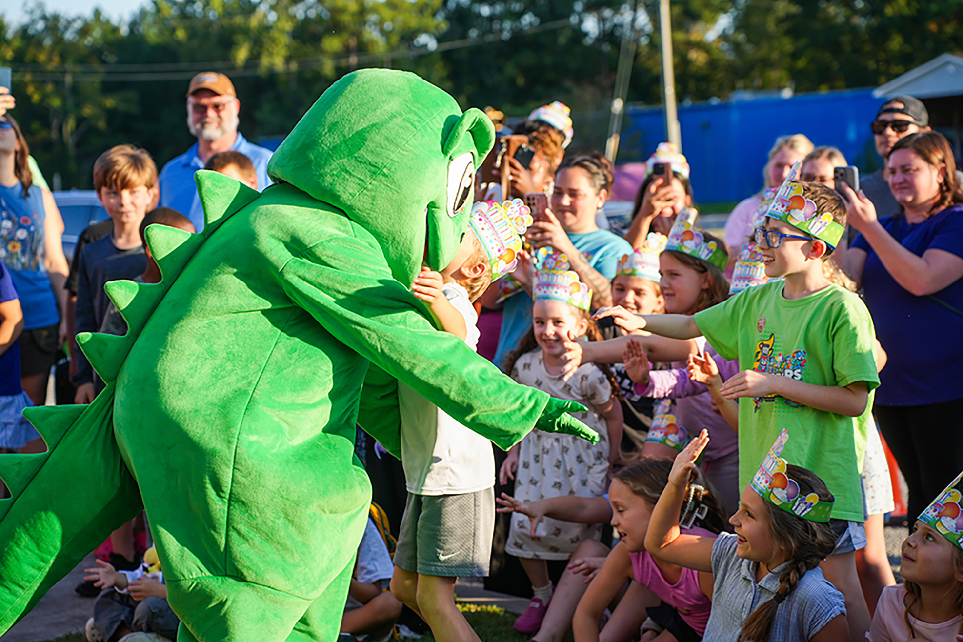 A mascot dressed as a green dinosaur interacts with children. Behind, a crowd of adults and children watch.