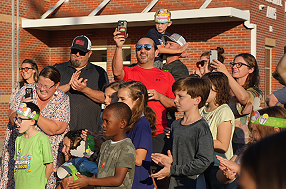 A group of people stands outside a brick building, some holding phones to capture the moment.