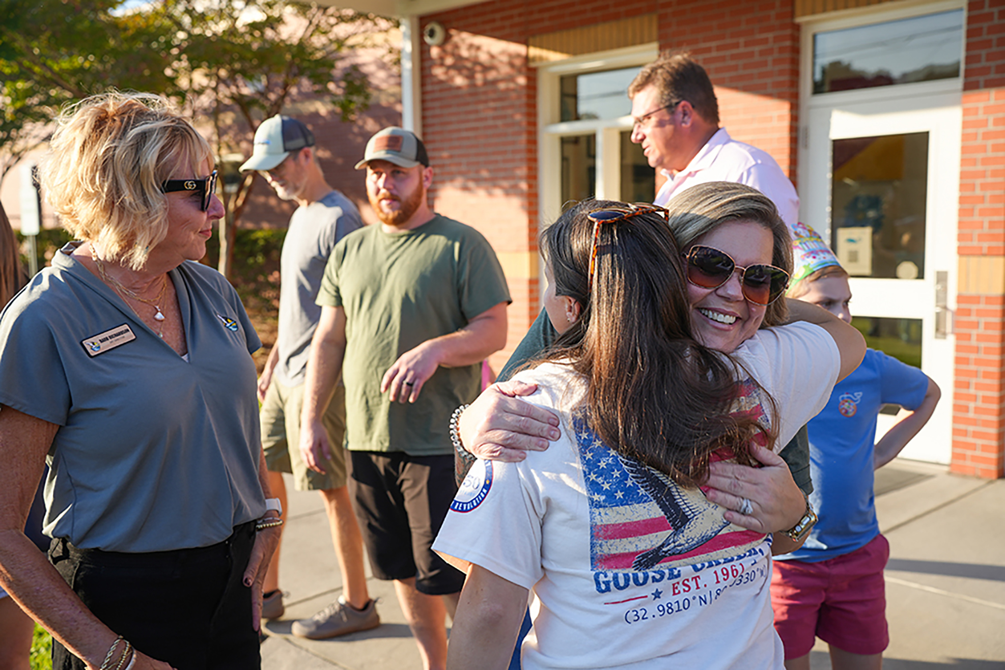 Adults stand outside a brick building, a woman hugs another woman. A man and a child stand behind them.