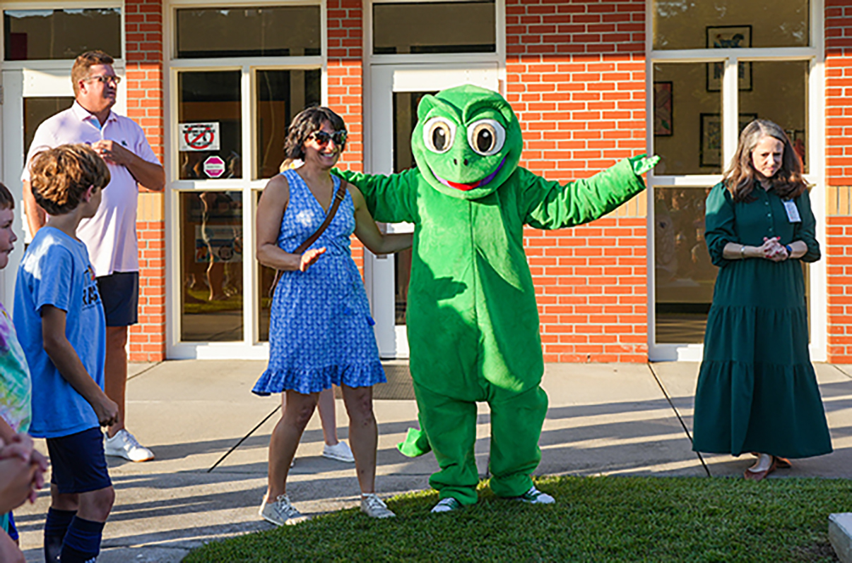 A group of people outside a building with glass doors. A person in a green mascot costume stands among them.