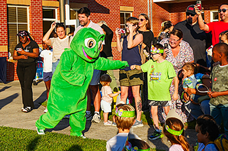 A mascot engages with children, while a crowd gathers outside a building. Adults and children are dressed casually.