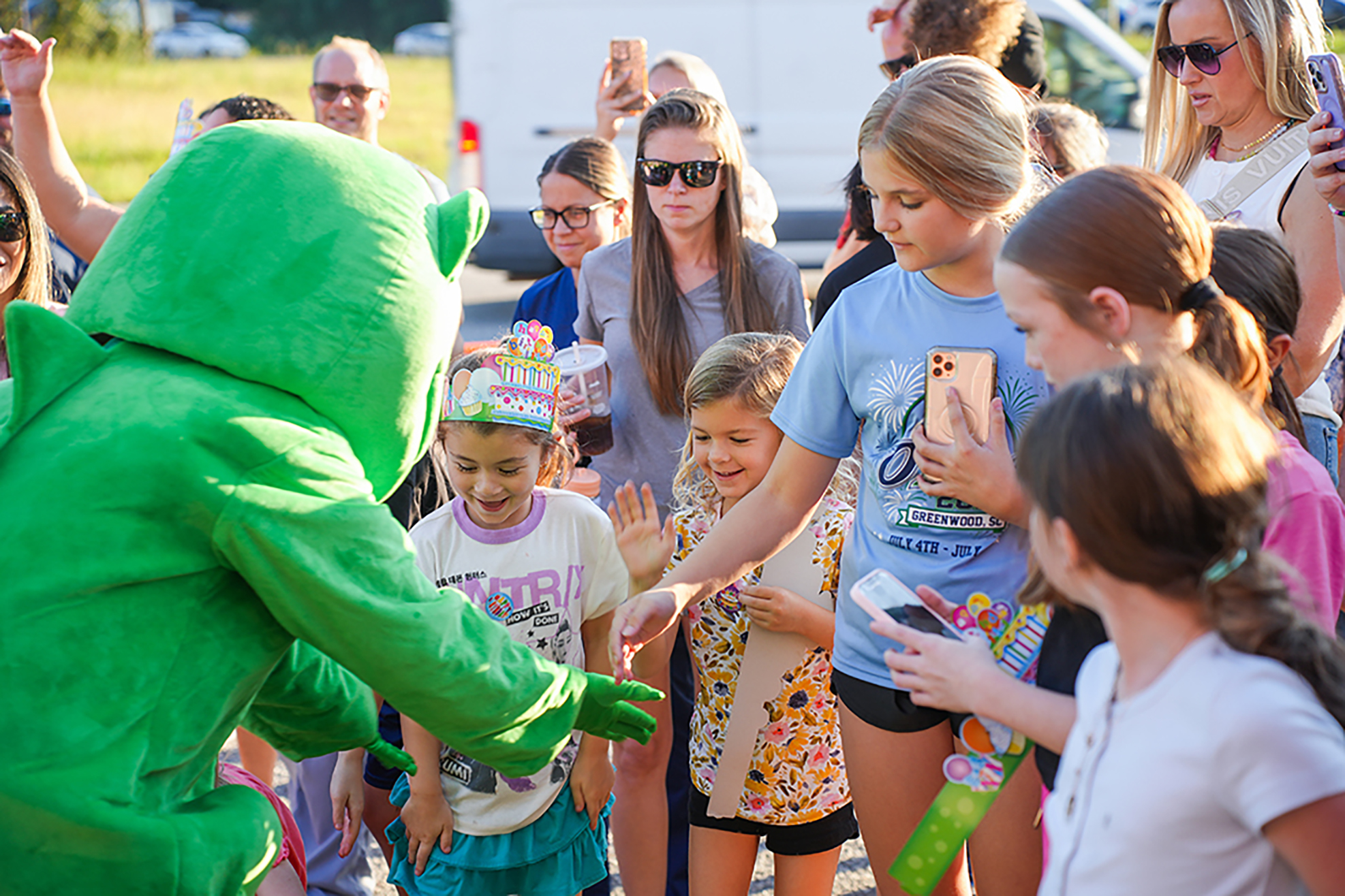 Green mascot in costume engages with children and adults in a crowd. The background shows a white van and grassy area.
