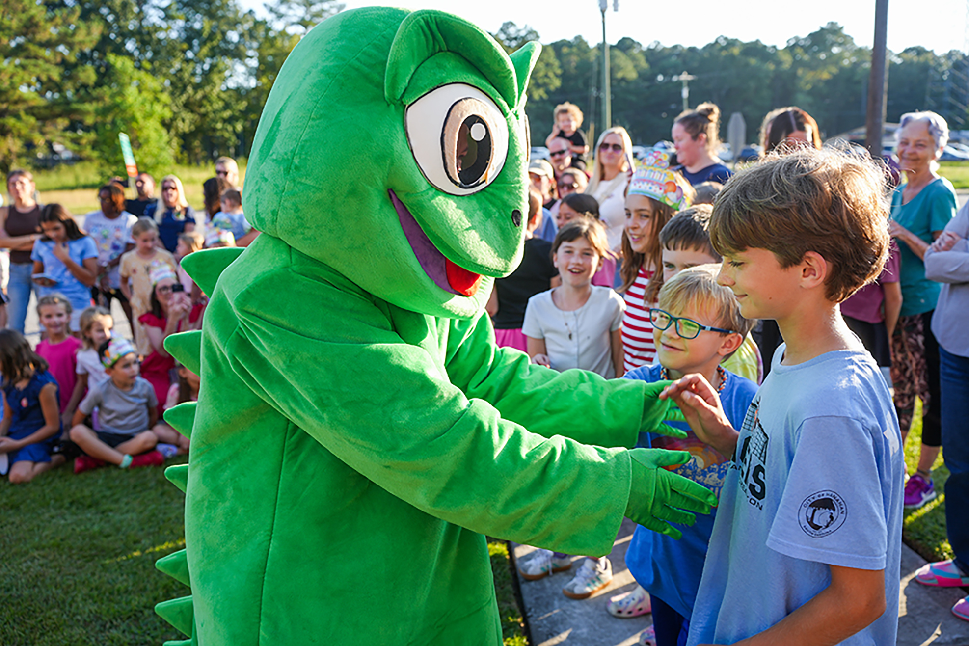 A green mascot in a costume interacts with two children in a grassy area. An audience of people is in the background.