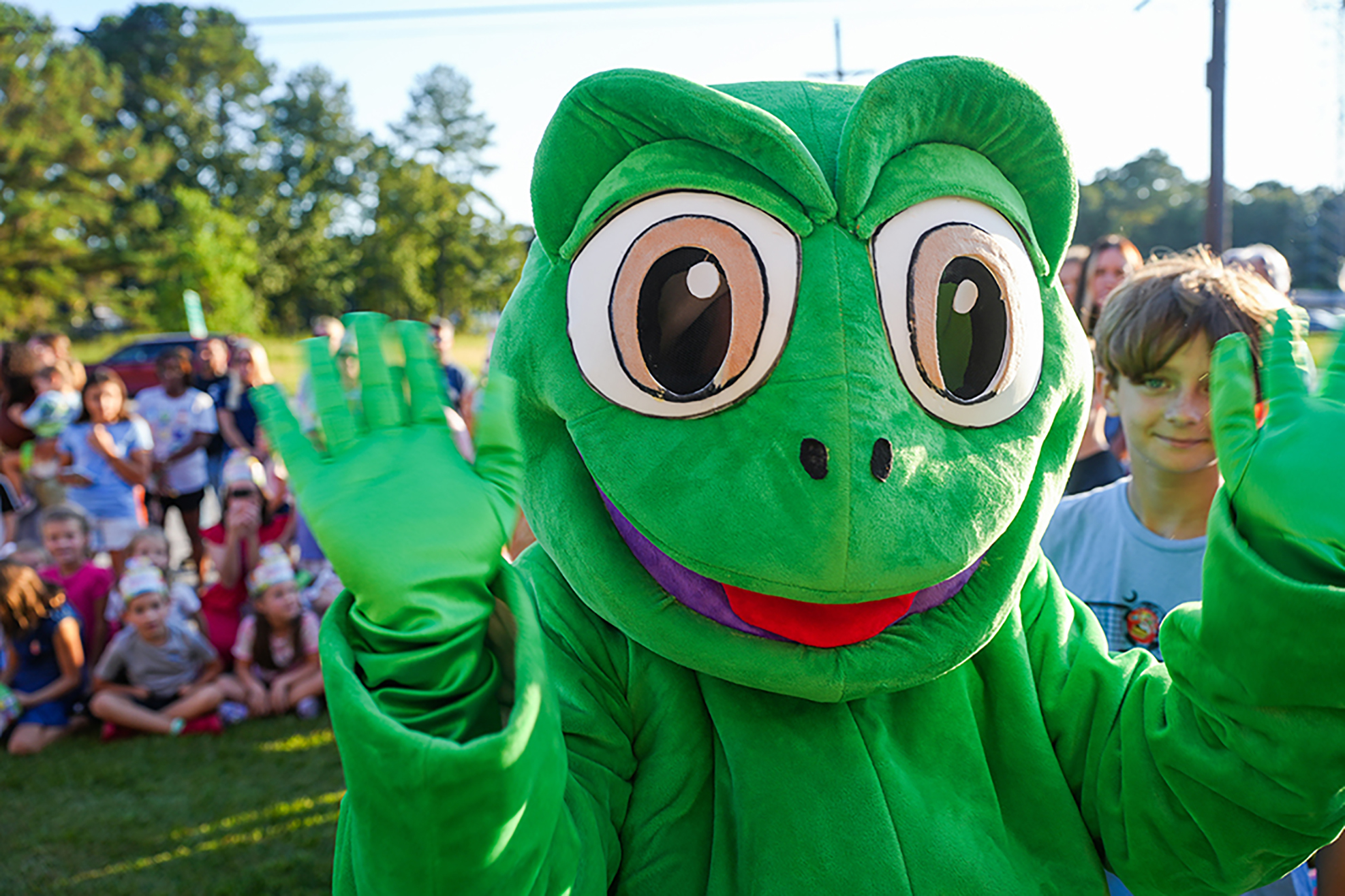 A frog mascot waves at a group of children sitting on the grass, with green trees in the background.