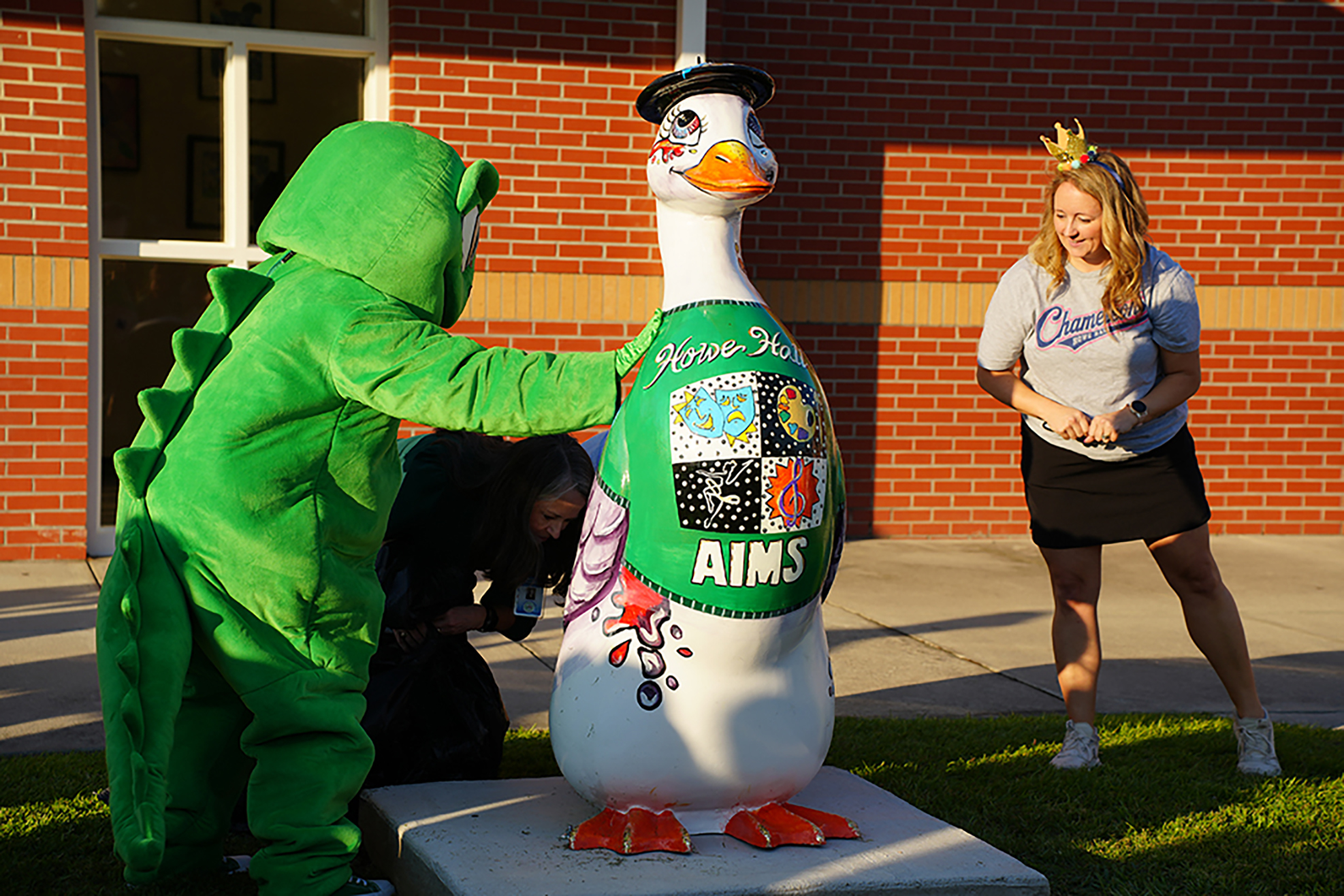 Person in mascot costume leans toward large bird statue while another stands by. Behind them is a brick building.