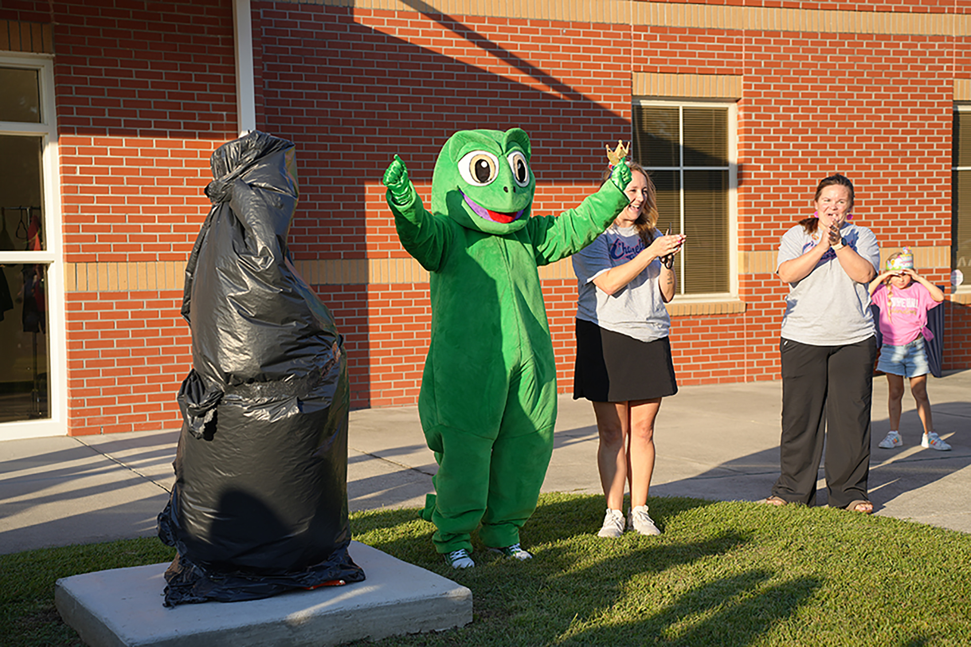 A person in a green mascot costume stands beside a black sculpture on grass. Behind them, two adults and two children stand and clap.