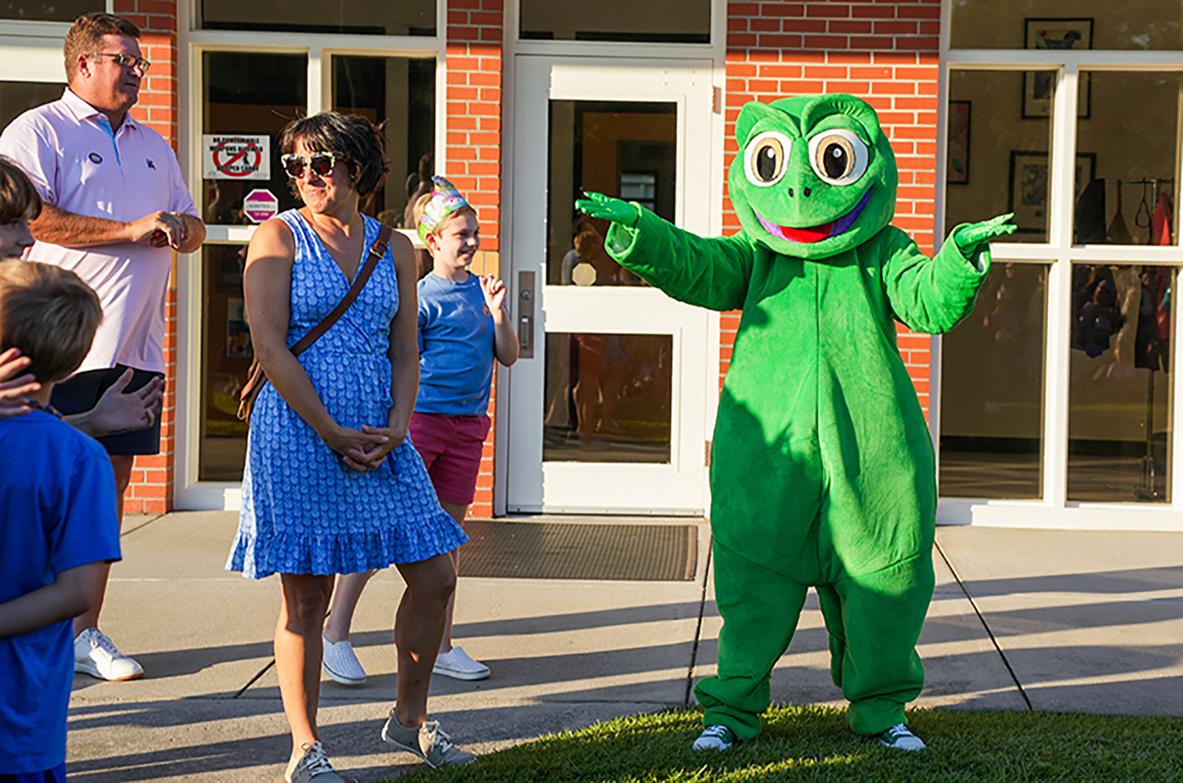A group of people in front of a building. A person in a frog mascot costume gestures to the side.