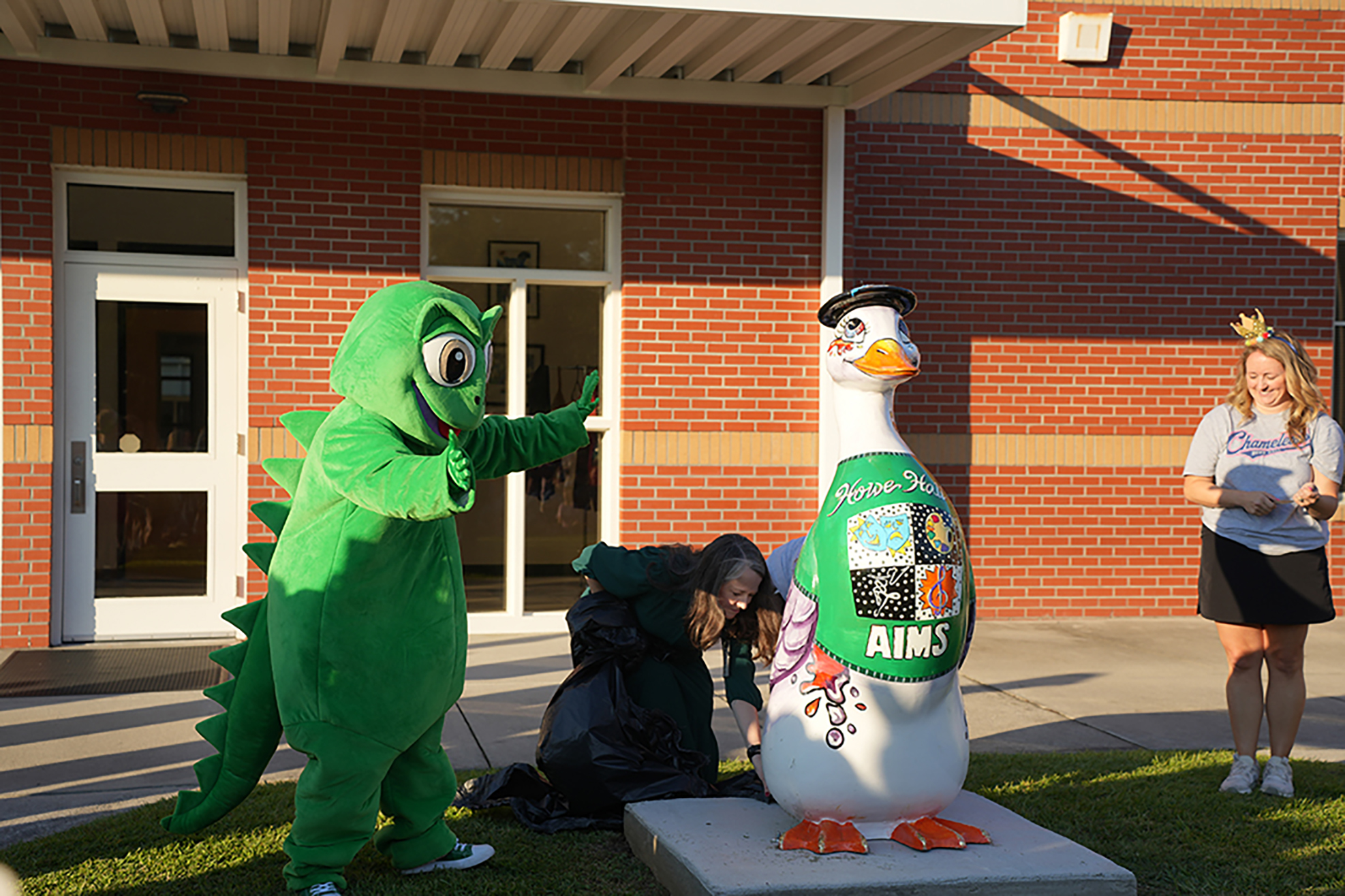 A person in a green dinosaur mascot costume waves. Another person is painting a duck statue. A girl in white sneakers watches.
