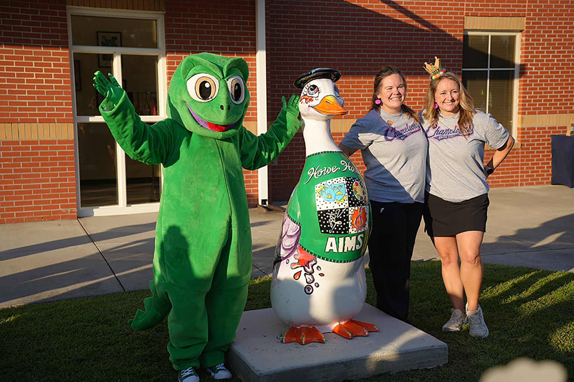 Three people stand in front of a brick building with a mascot on grass. The mascot is green.