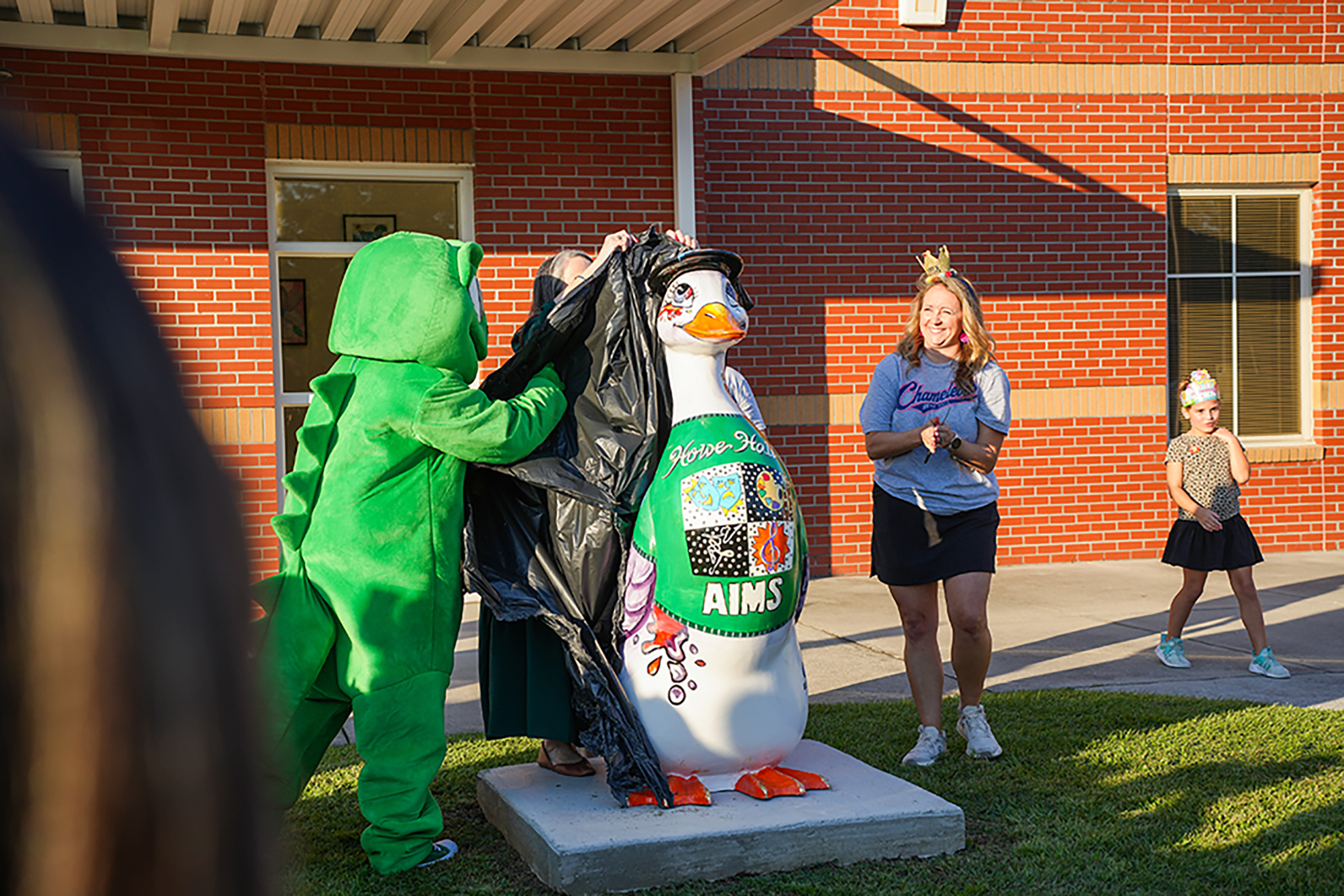 People in costumes, one a dinosaur, one a penguin, and another a child, in front of a brick building.
