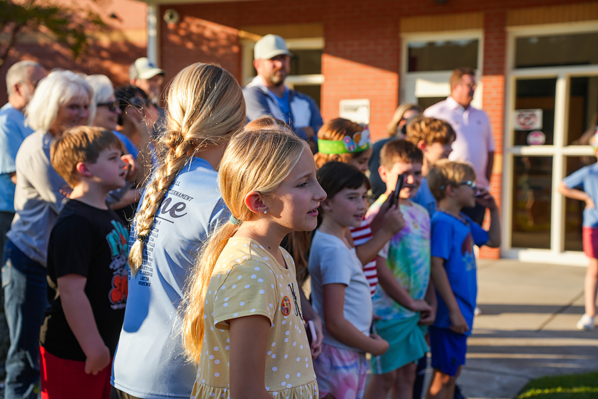 Several children and adults gather in front of a brick building, some wearing hats and glasses.