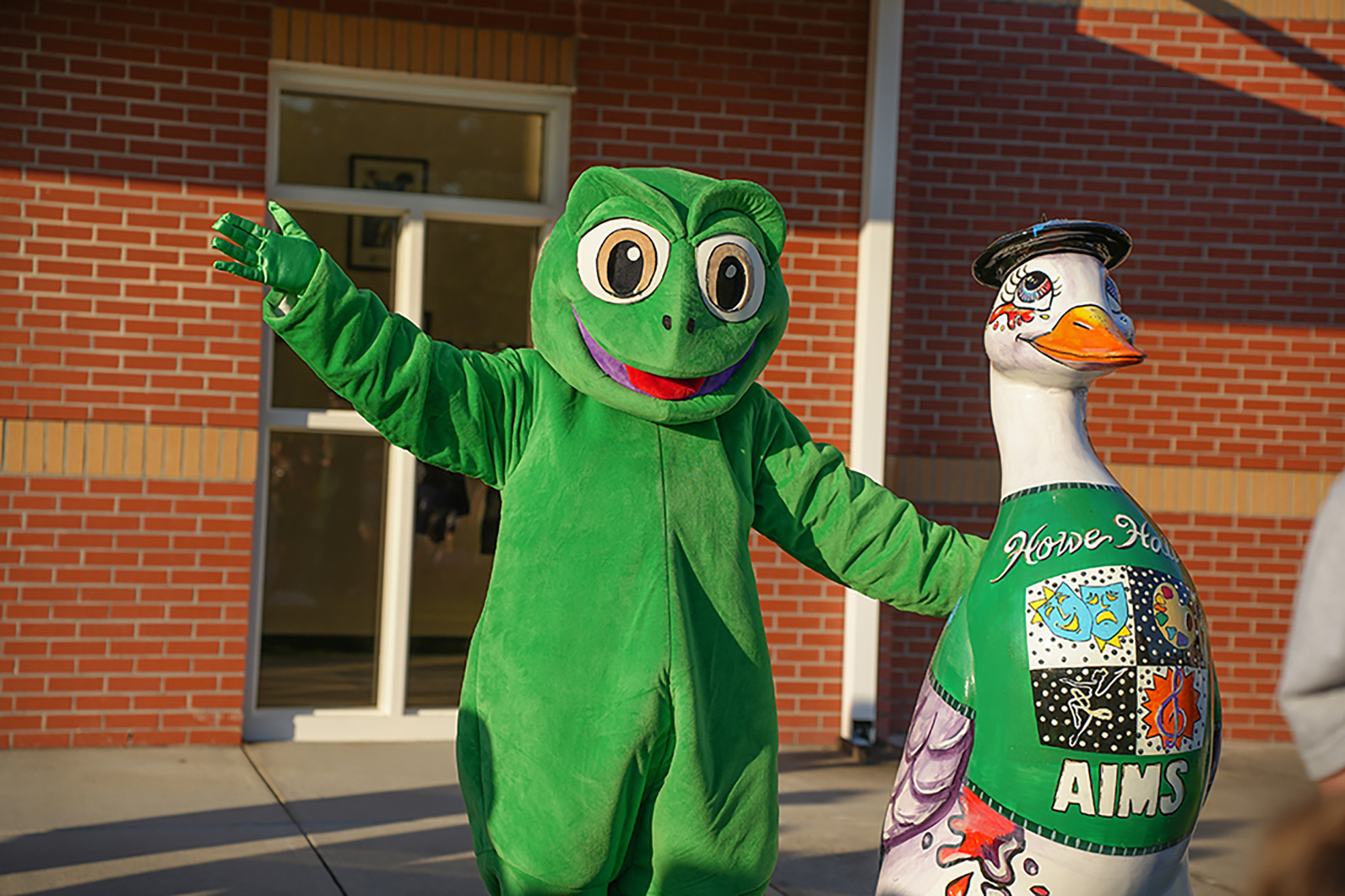 Two mascots in front of a building. One is a green frog mascot in a green costume. The other is a white goose.