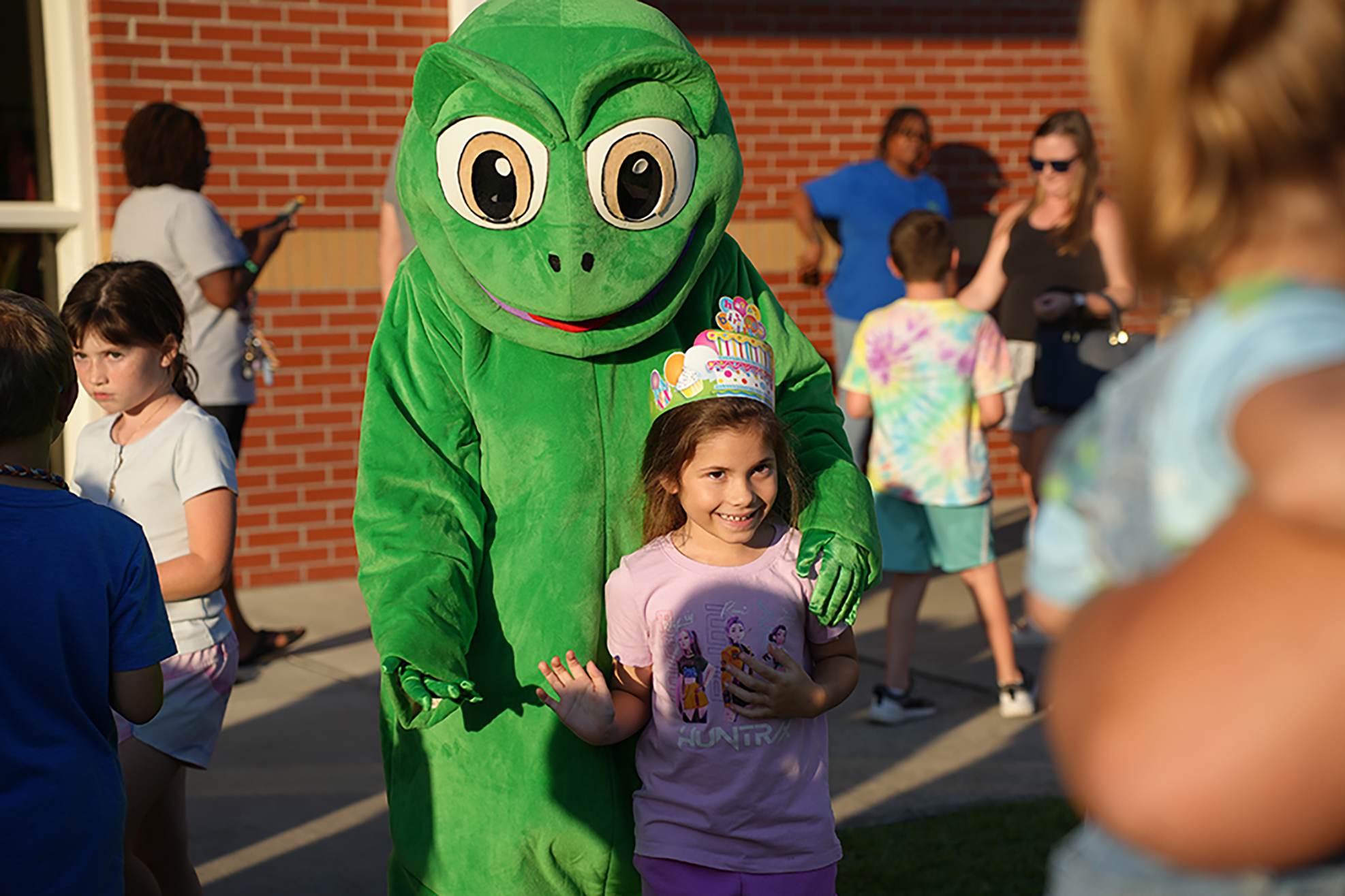 A green mascot with large eyes stands with a young girl in a pink shirt, surrounded by children in casual clothes.