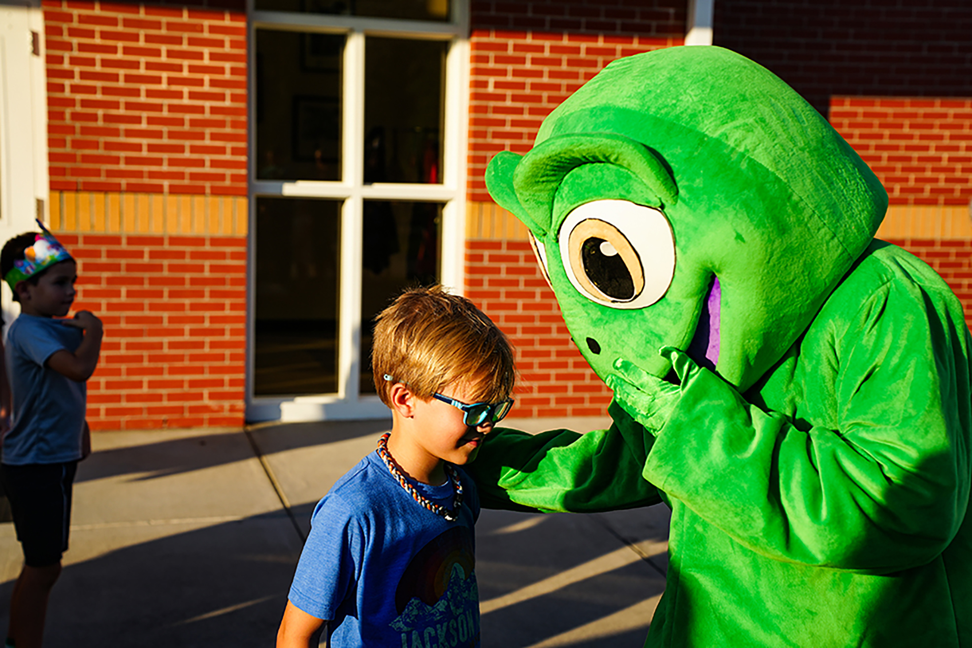 A child wearing glasses stands next to a large green mascot in front of a red brick building.
