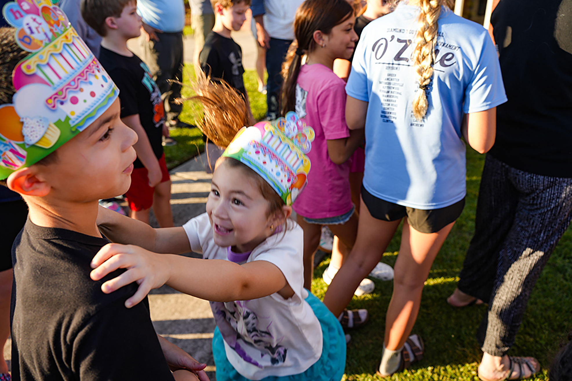 A child with a birthday hat looks at another child. Behind them, adults and children gather on a grassy area.