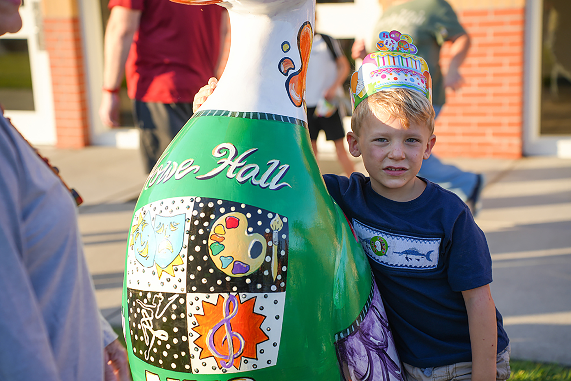 A child stands beside a large, colorful sculpture of a bottle. The sculpture has various images on it.